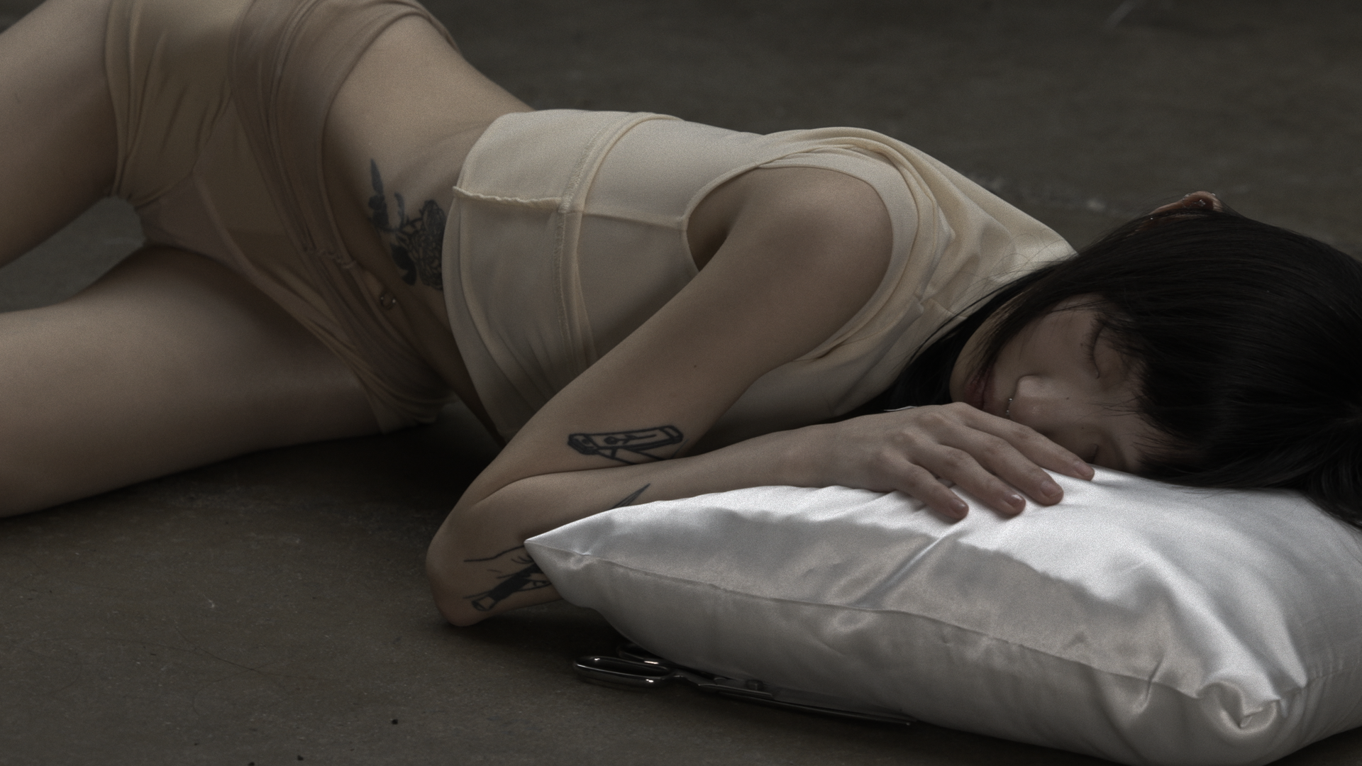 Person lying on concrete floor with head resting on a white pillow, eyes closed, body in nude-toned clothing; scissors placed beneath pillow edge, soft lighting throughout.