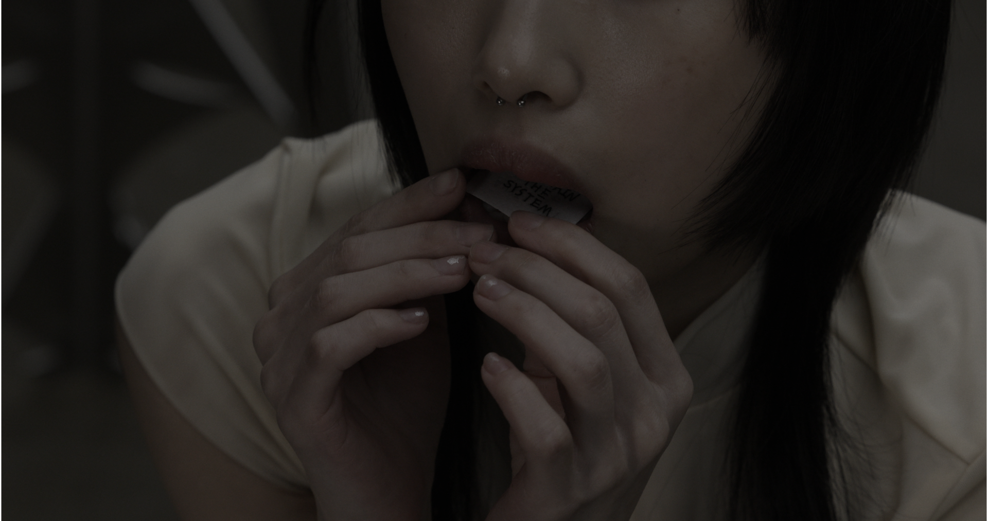 Close-up of mouth holding a small paper labeled “MAINTAIN THE SYSTEM,” fingers pressing it inward; neutral tones, hair framing face, expression controlled.