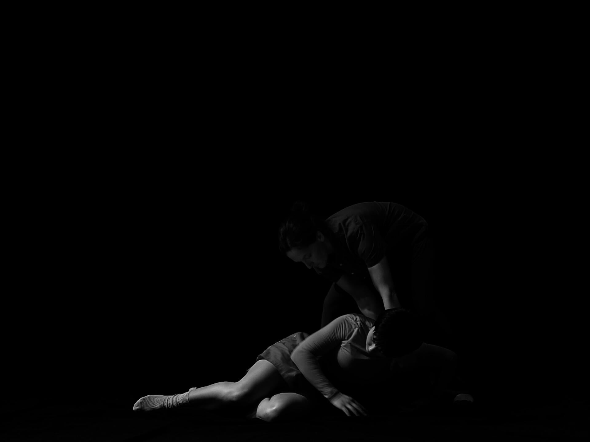 Two (female/non-binary identifying) figures are pictured against a black background in this black and white video still. 