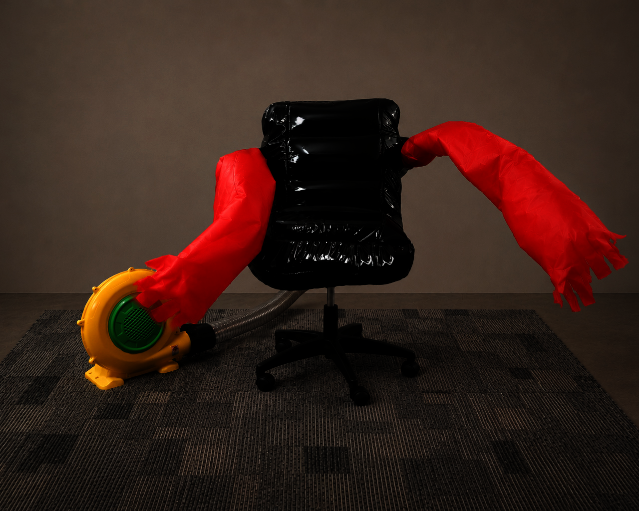 Front shot of the CHAIR FOR WORKPLACE MORALE with a neon yellow and green air blower, vinyl inflatable office chair, and red inflatable tube arms swinging side to side.