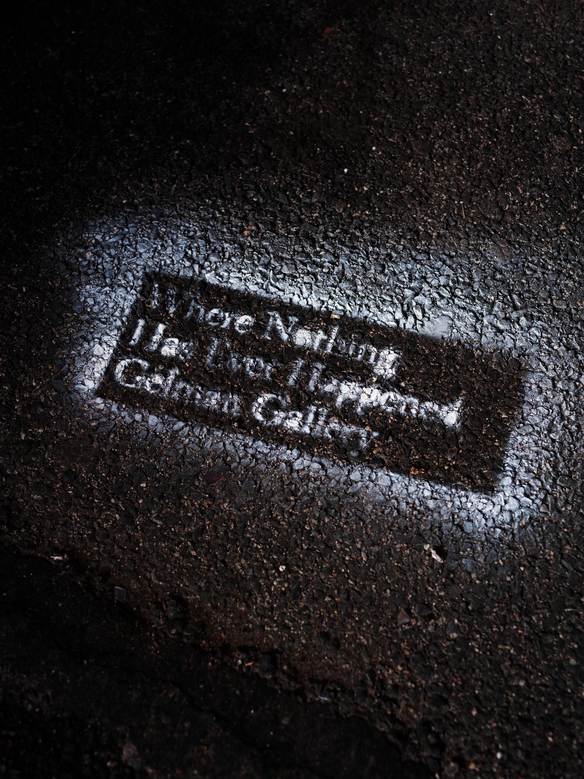 Close-up of a white stenciled text on dark, wet asphalt reading "Where Nothing Has Ever Happened / Gelman Gallery," photographed at an angle to emphasize the rough texture of the pavement.