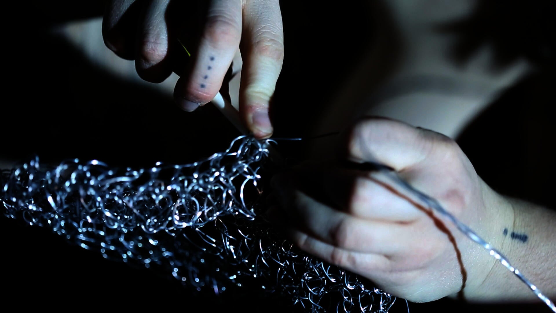 Close-up of hands crocheting thin silver wire into a tangled mesh, with one holding the hook and the other threading wire. Dramatic and focused lighting highlights metallic texture.