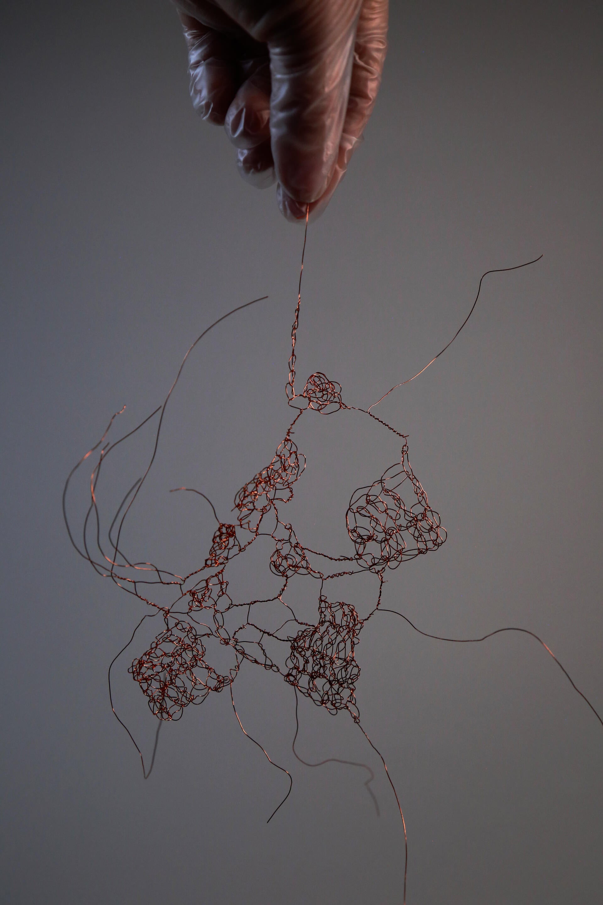 Gloved hand holds delicate copper wire sculpture, forming tangled, organic shapes suspended against a white background.