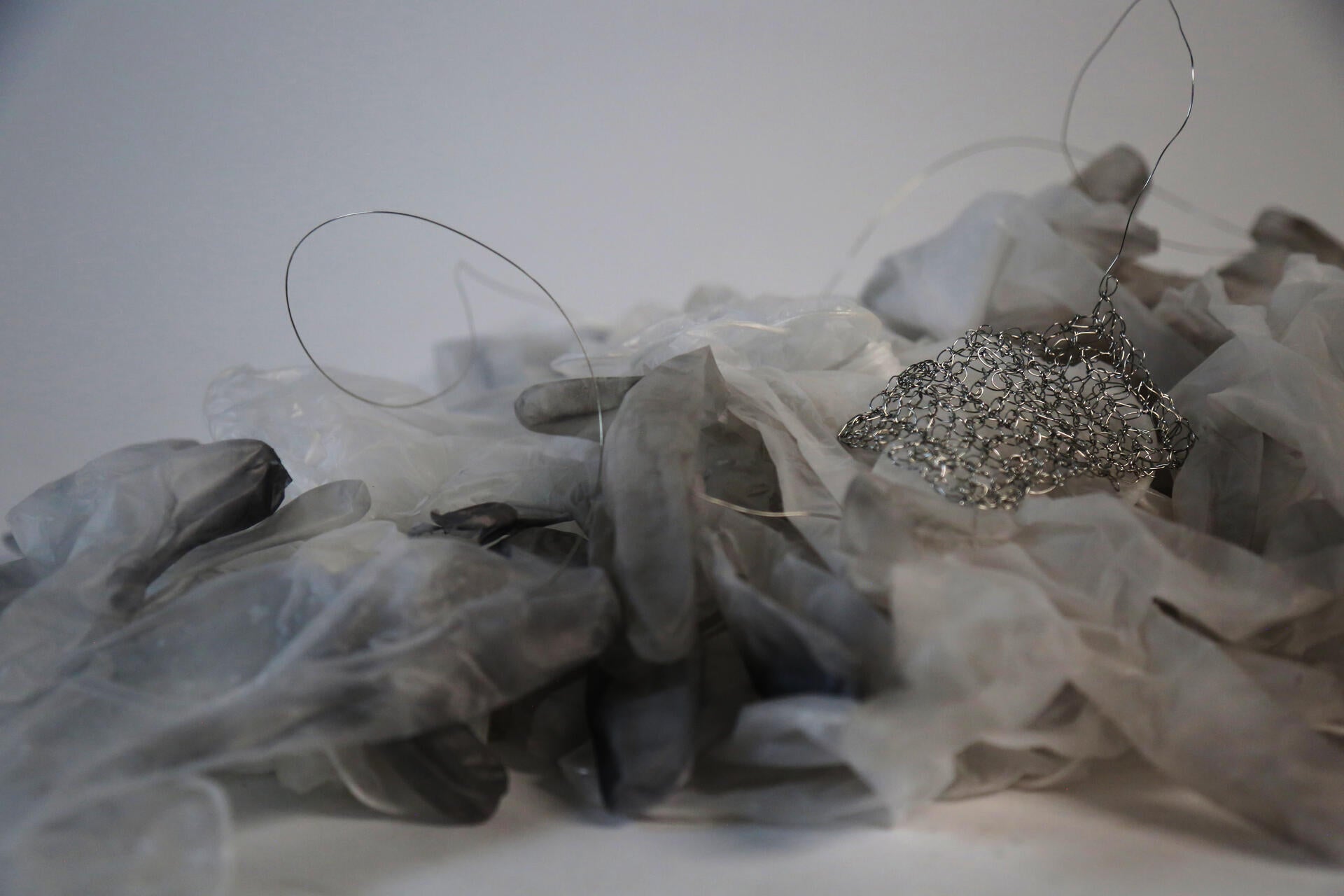 A pile of semi opaque used vinyl gloves with a delicate crocheted piece made from thin silver wire placed amidst the pile.