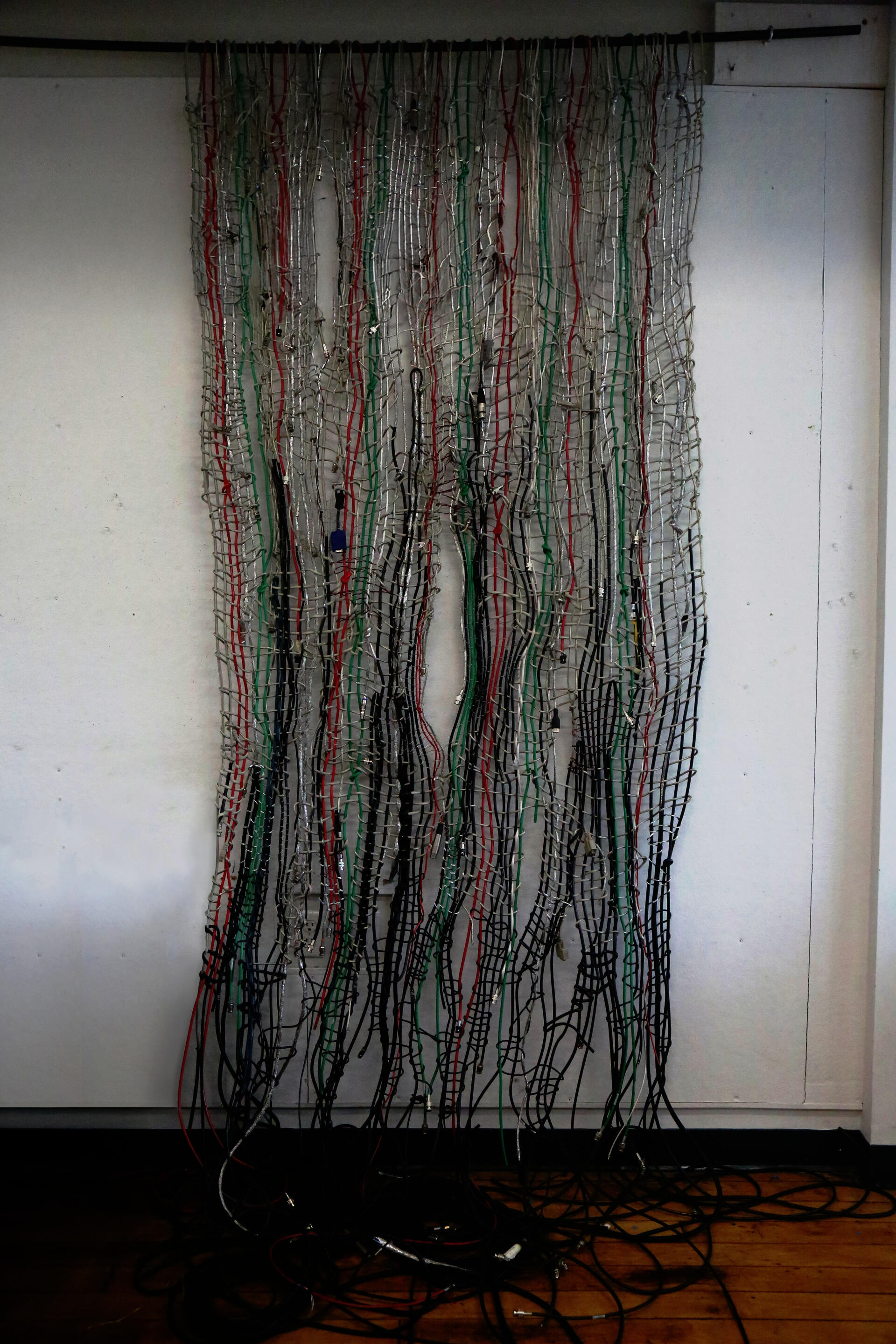 Large, dense textile of multicolored cables hanging vertically, woven with stripped metal wires against a light background, creating a chaotic, net-like pattern.