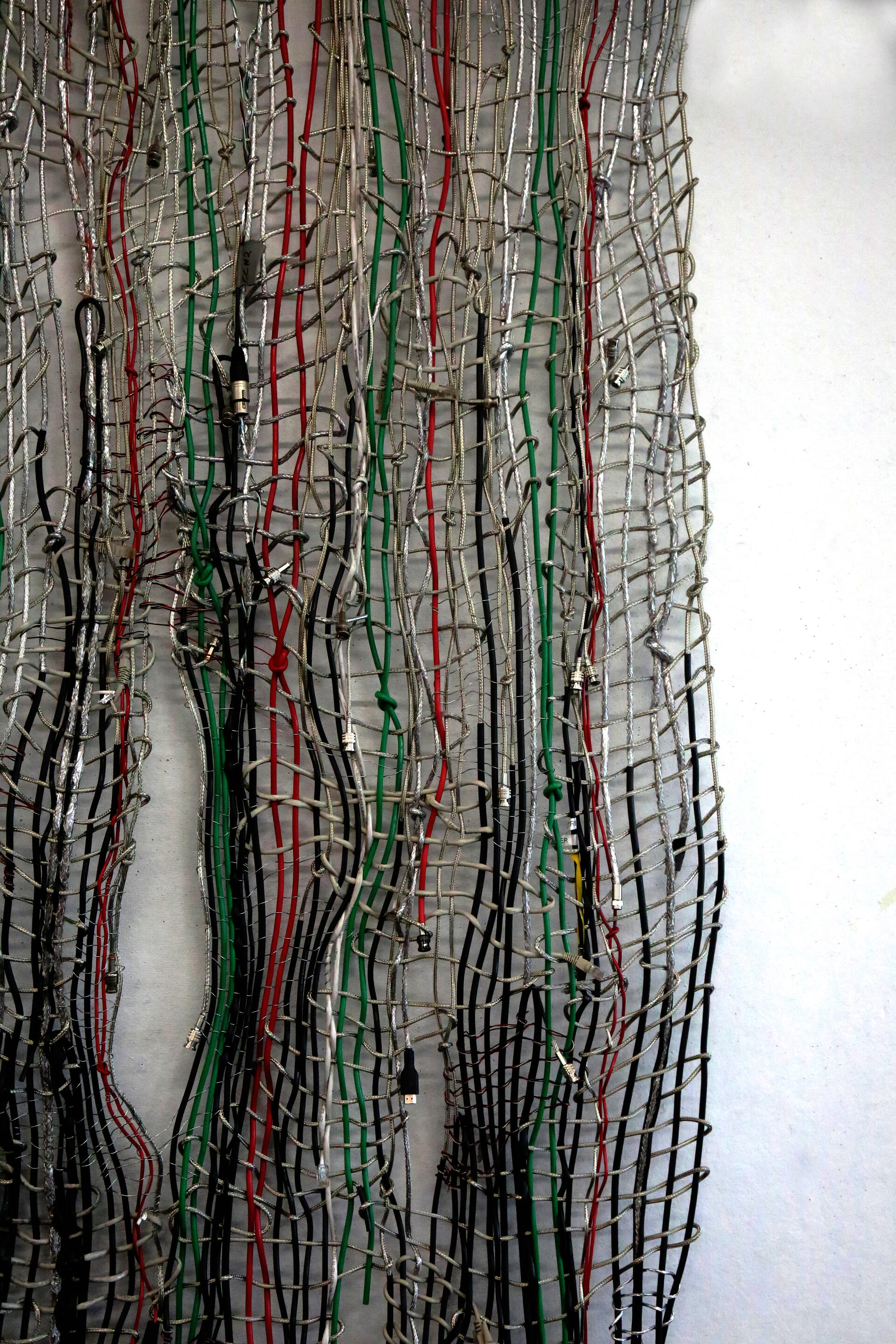 Vertical off center close up of weave made from various cables with the red, green, and black cables highlighted against the other stripped silver wires against a white background.