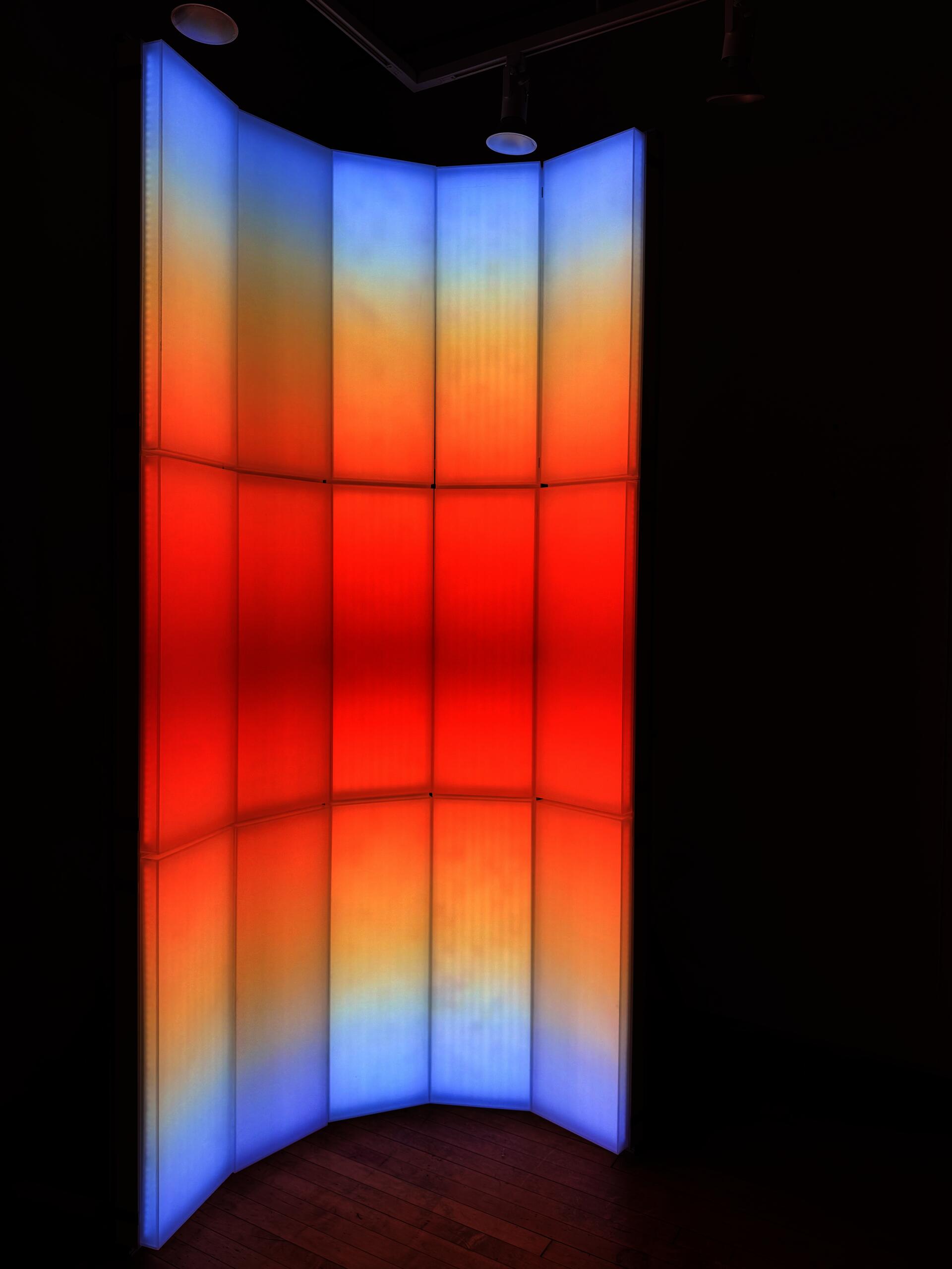 A light-based installation composed of vertical translucent panels, with blue, orange, and red gradients that evoke the color transition of sunrise and sunset.