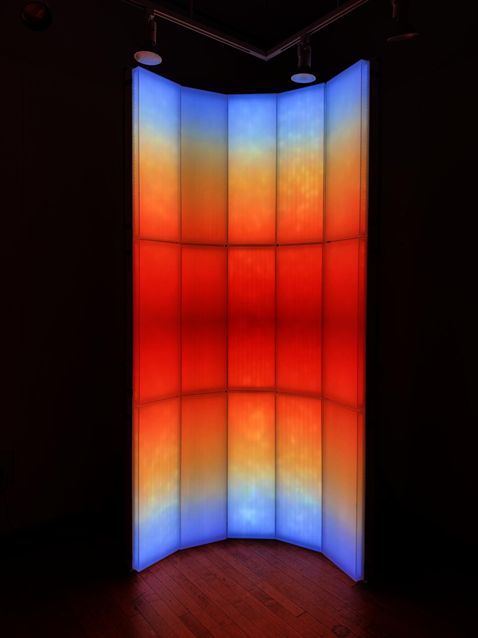 A light-based installation composed of vertical translucent panels, with blue, orange, and red gradients that evoke the color transition of sunrise and sunset.