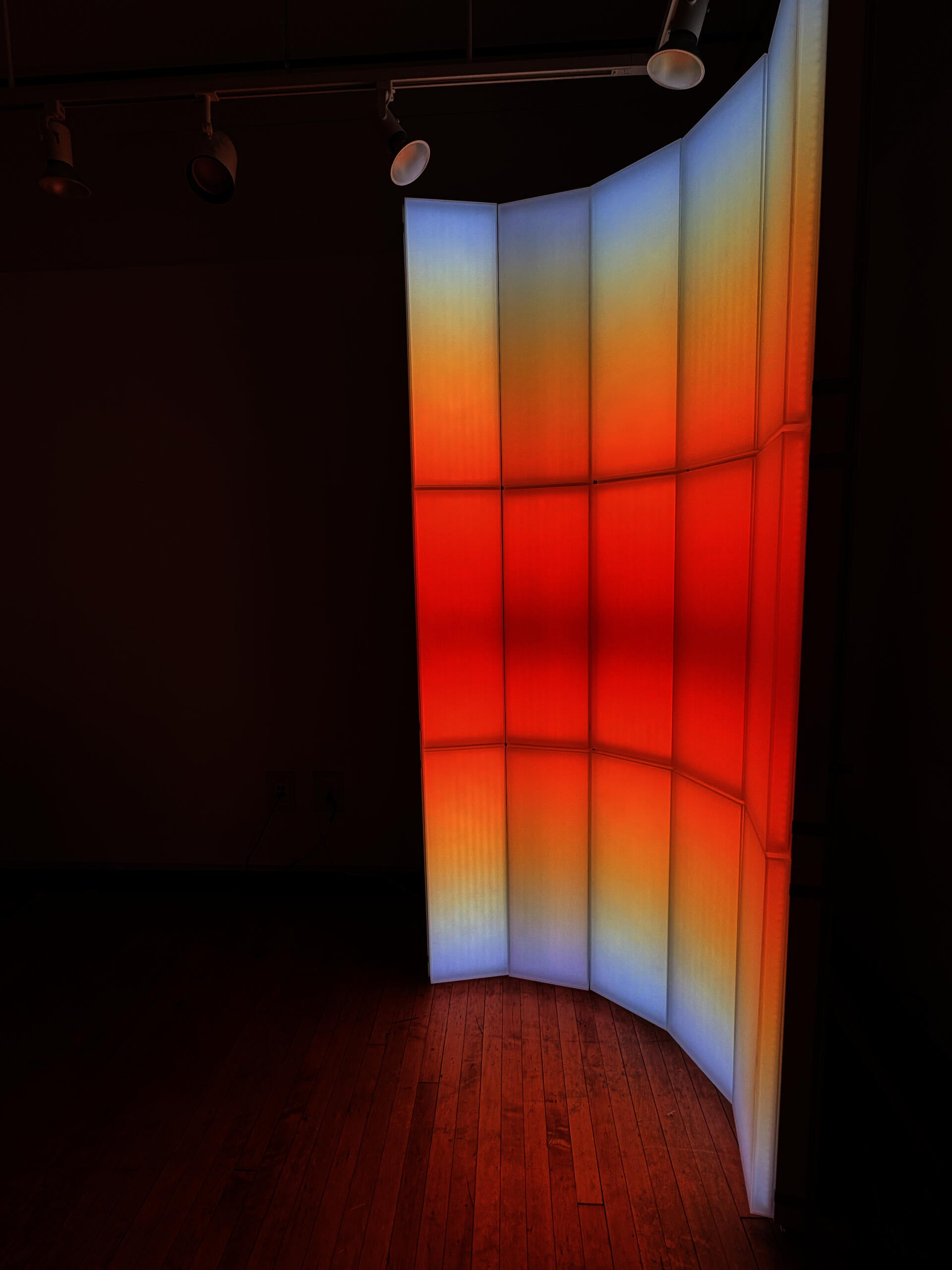 A light-based installation composed of vertical translucent panels, with blue, orange, and red gradients that evoke the color transition of sunrise and sunset.