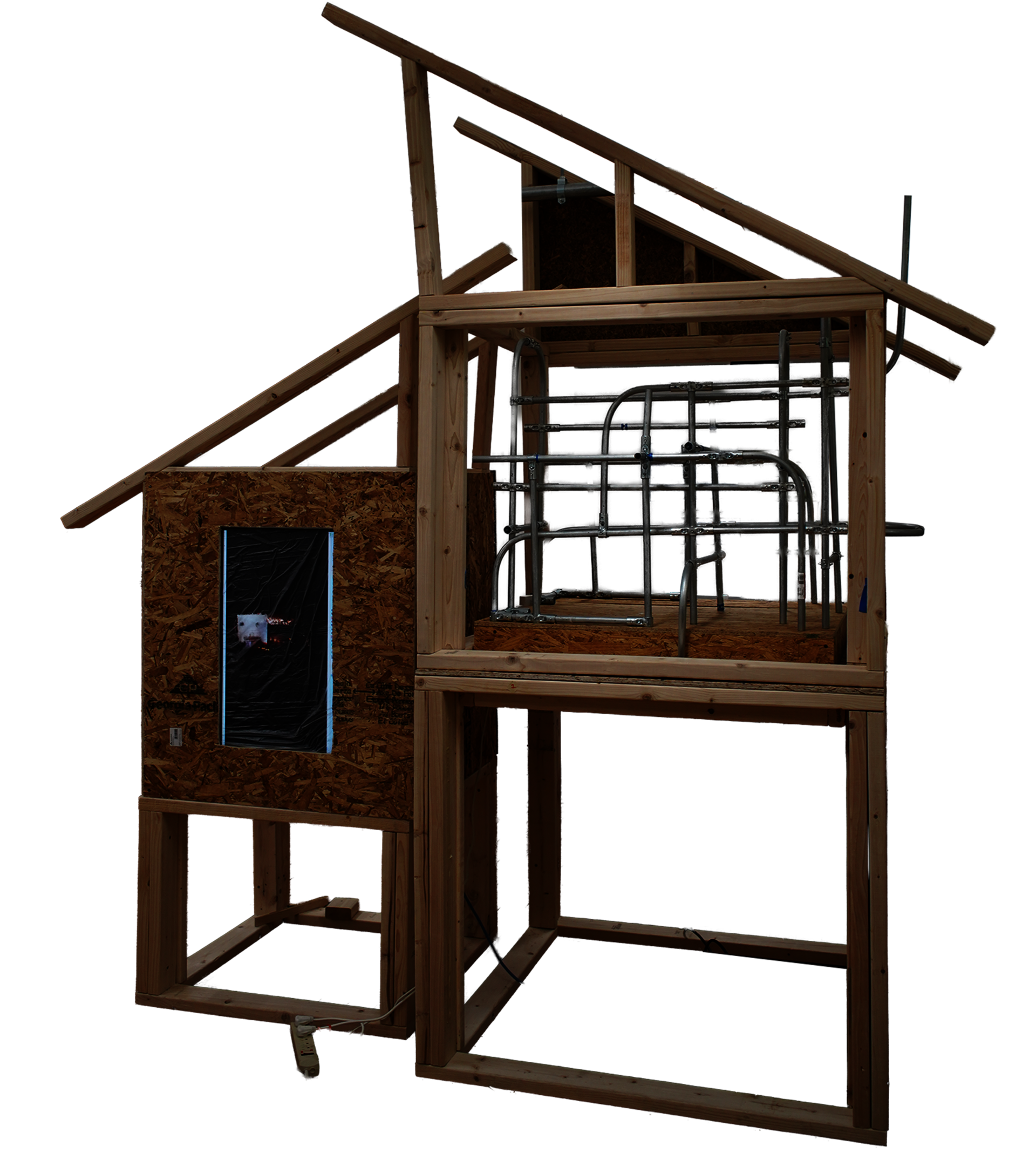 A multi-level architectural installation built from exposed raw lumber, resembling an unfinished house frame. The right side features an upper level densely filled with a geometric network of intersecting, bent metal conduits. 