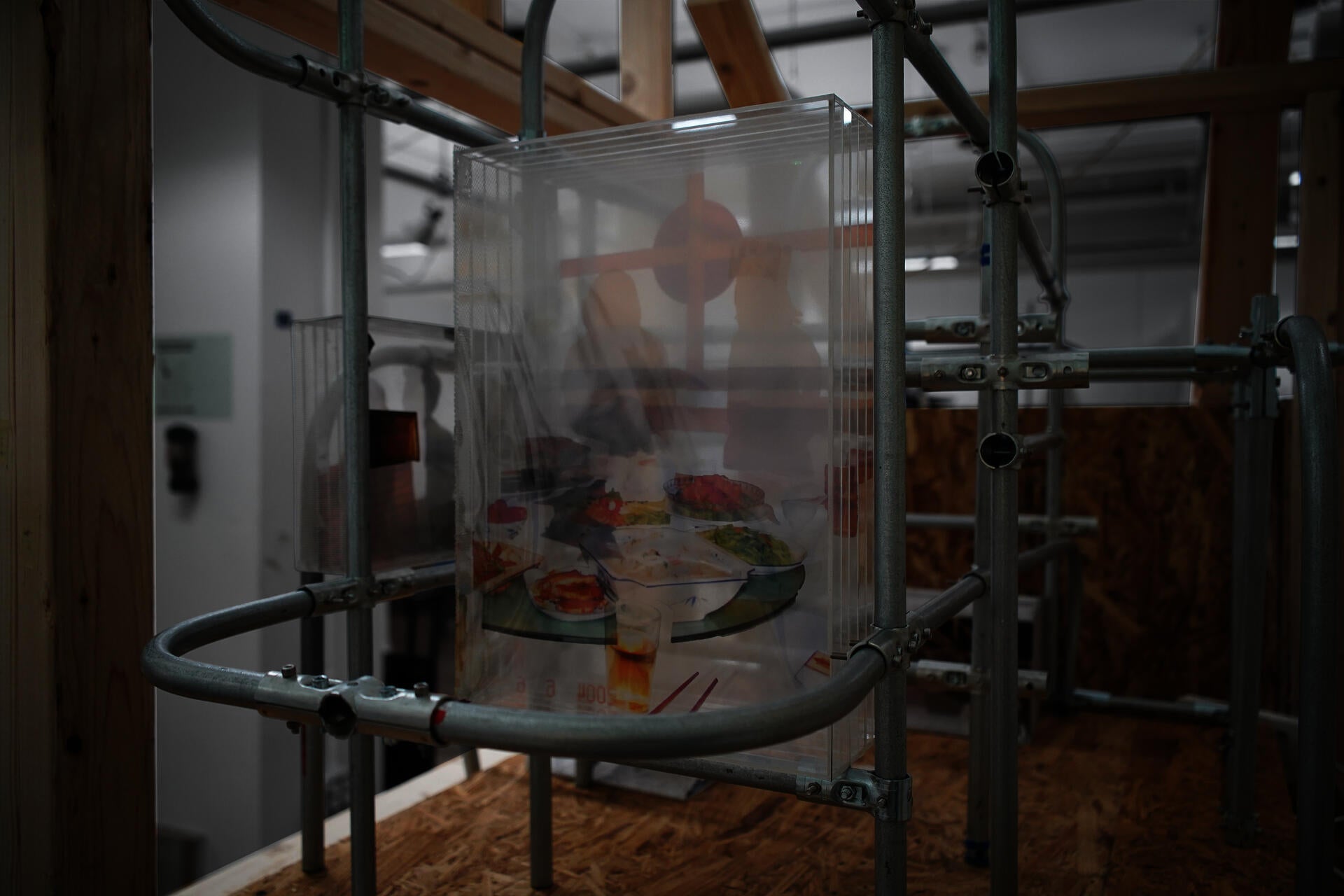 A detailed close-up of the art installation frame built from raw lumber and curved metal conduits. A multi-layered transparent acrylic box, held within the structure, contains a deconstructed archival photograph of a dinner table scene. 