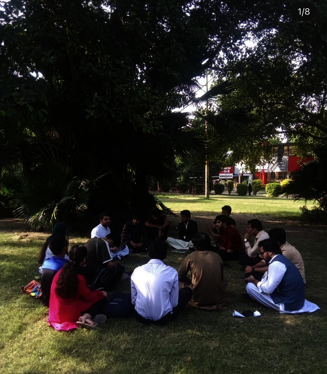 A circle of students and workers seated on a lawn in dappled sunlight, leaning toward each other in conversatiom; showcases knowledge shared without hierarchy