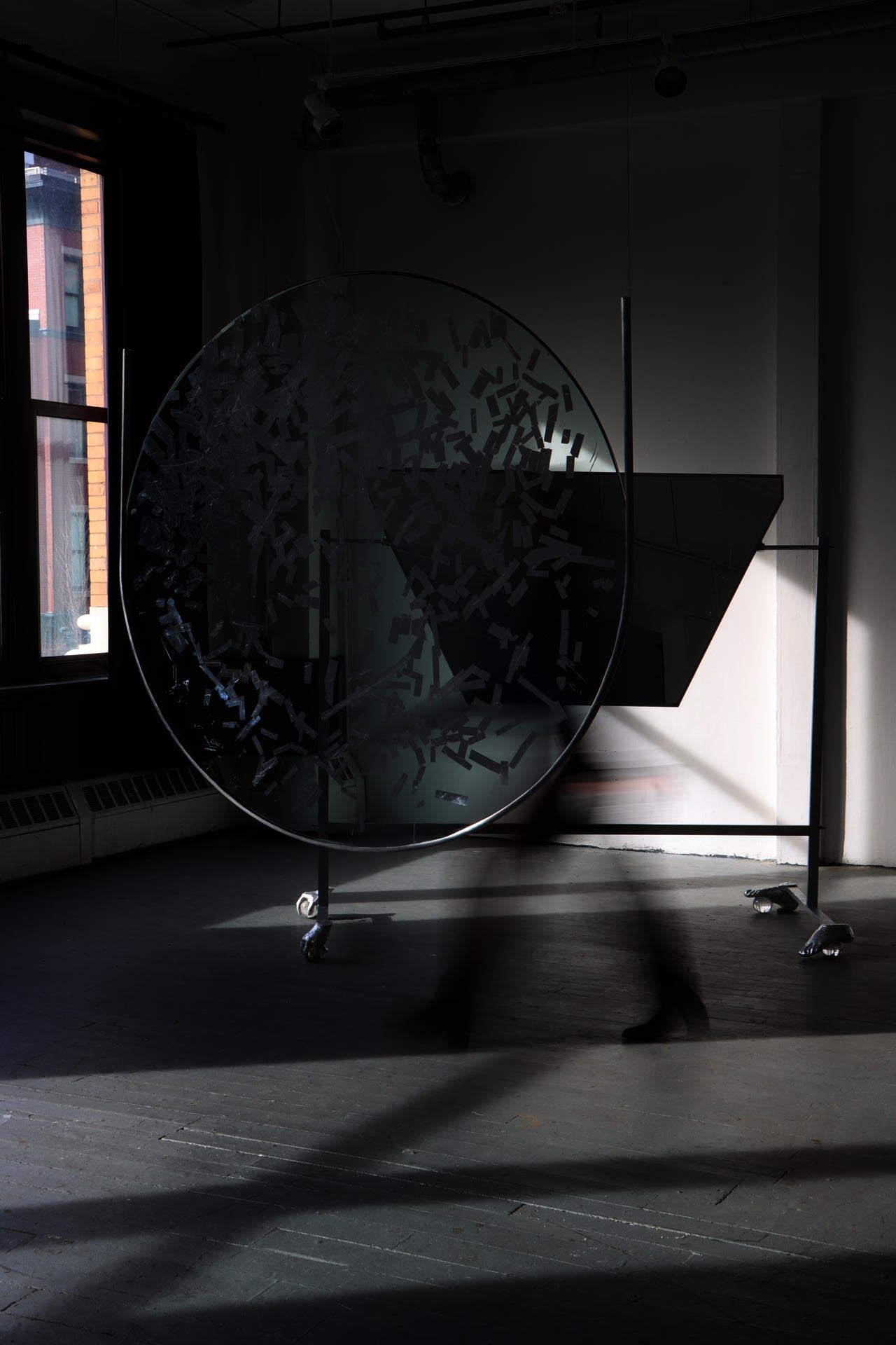  4ft diameter piece of glass hands supsended in a room, with part of its surfaced gilded silver. A person in motion passes behind it. Behind them is large piece of mirror angled on a aluminum stand, with feet made of hands and feet on crystal balls. 