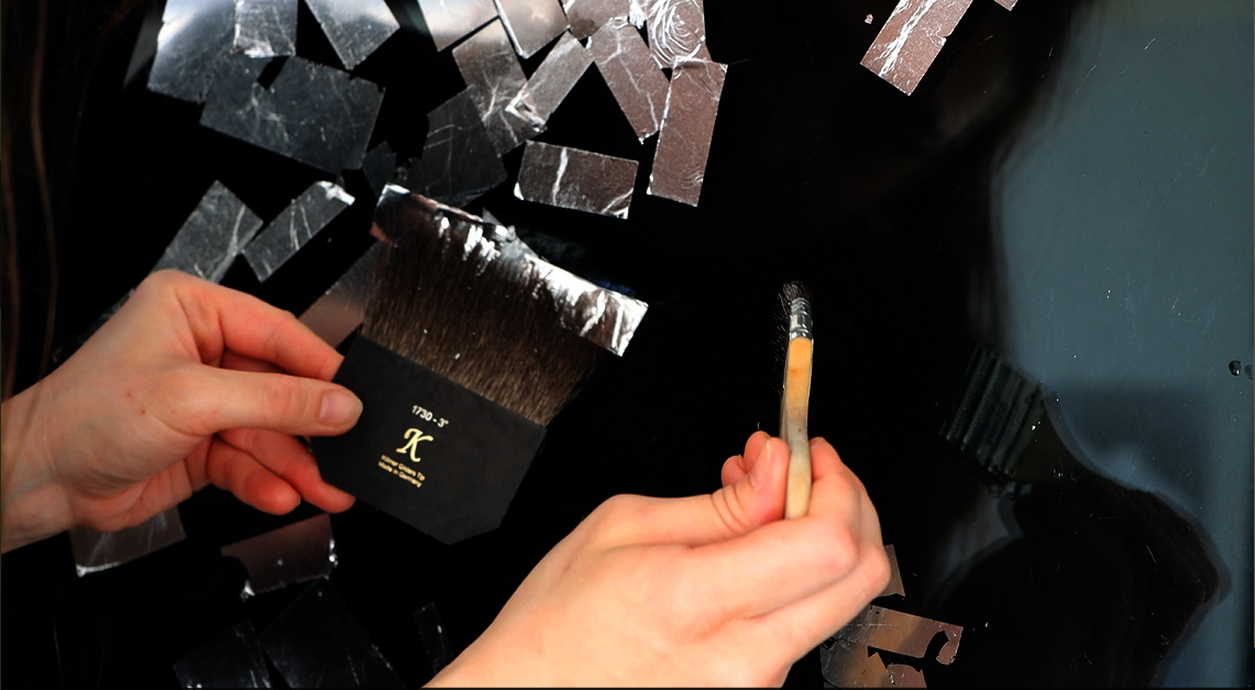 A close up shot of a person applying aluminum leaf to a glass with a brush for the leaf in one hand, and a brush for size in the other. The glass is against a black  background and the veins and wrinkles of the metal contrast with the smoothness of the glass surface.