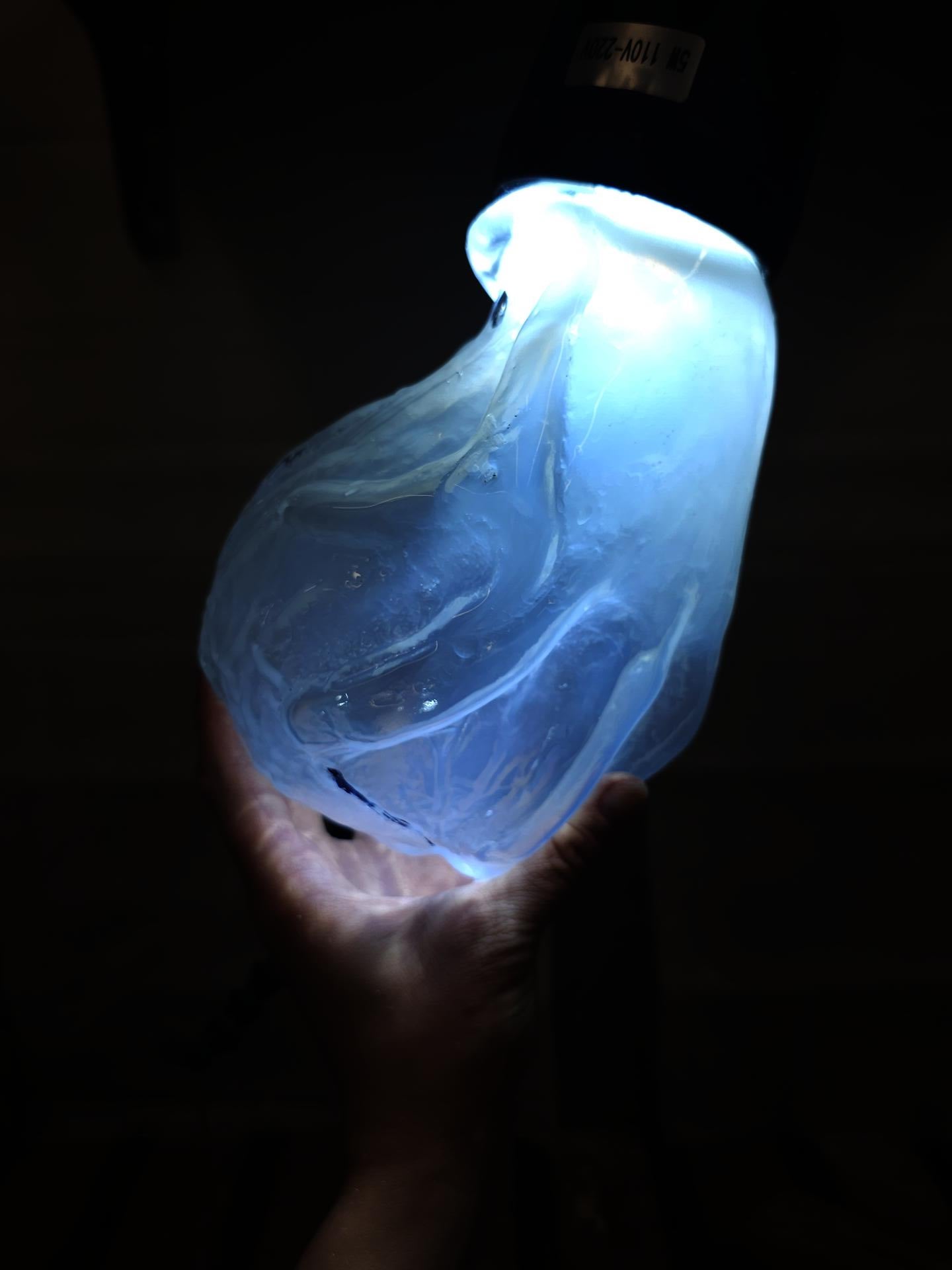 Against a darkened background, a pale hand holes up a piece of blown glass the shape of a hand grasping a sphere-ish shape, glowing blue from within. 