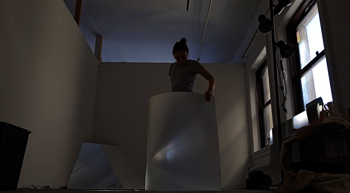 A woman stands in the middle of a sunlight room in waning light. She wears a t-shirt and has brown hair in a bun. She holds a giant fresnel lens around her body, looking down to see just reflected light and the disappearance of her body. 