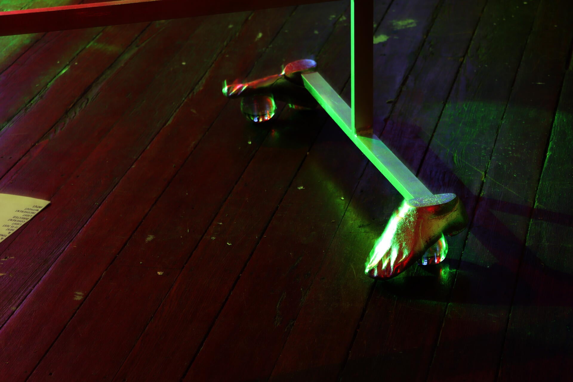 Close up of a silver sculpture base, whose feet are aluminum feet in jazz shoes, over solid glass balls. The light is green from one direction, and red from another, fuzzy and a little out of focus. 