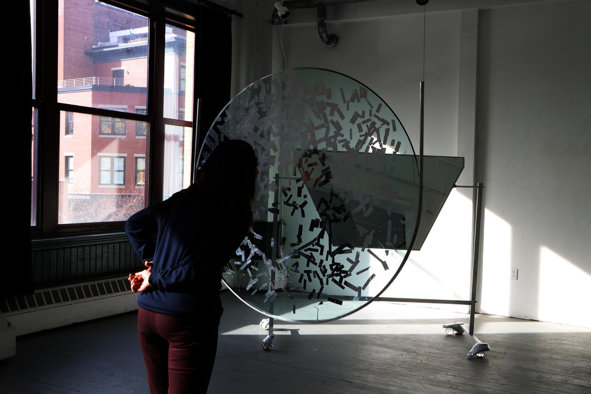4ft diameter piece of glass hands supsended in a room, with part of its surfaced gilded silver. A person in motion passes behind it. Behind them is large piece of mirror angled on a aluminum stand, with feet made of hands and feet on crystal balls.