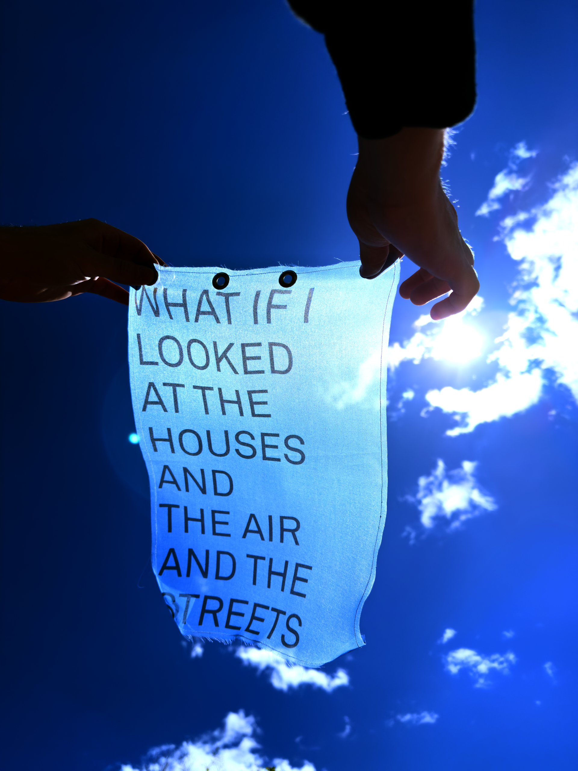 Someone holding a blue fabric print up to the sky that reads "what if I looked at the houses, and the air, and the streets