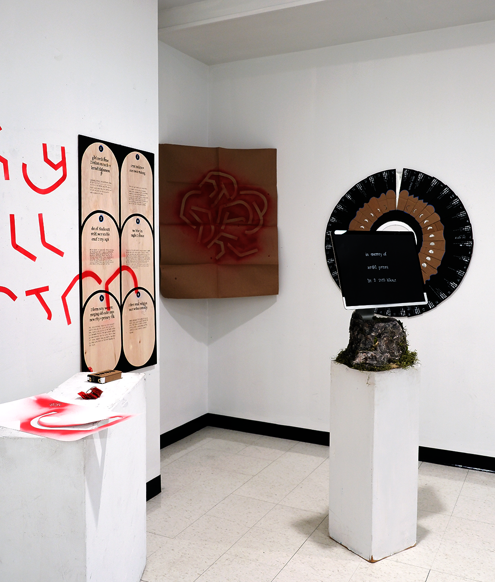 An installation critical of AI, showing an iMac within a sculpted rock on a pedestal, various red, black, and tan typographic objects on walls