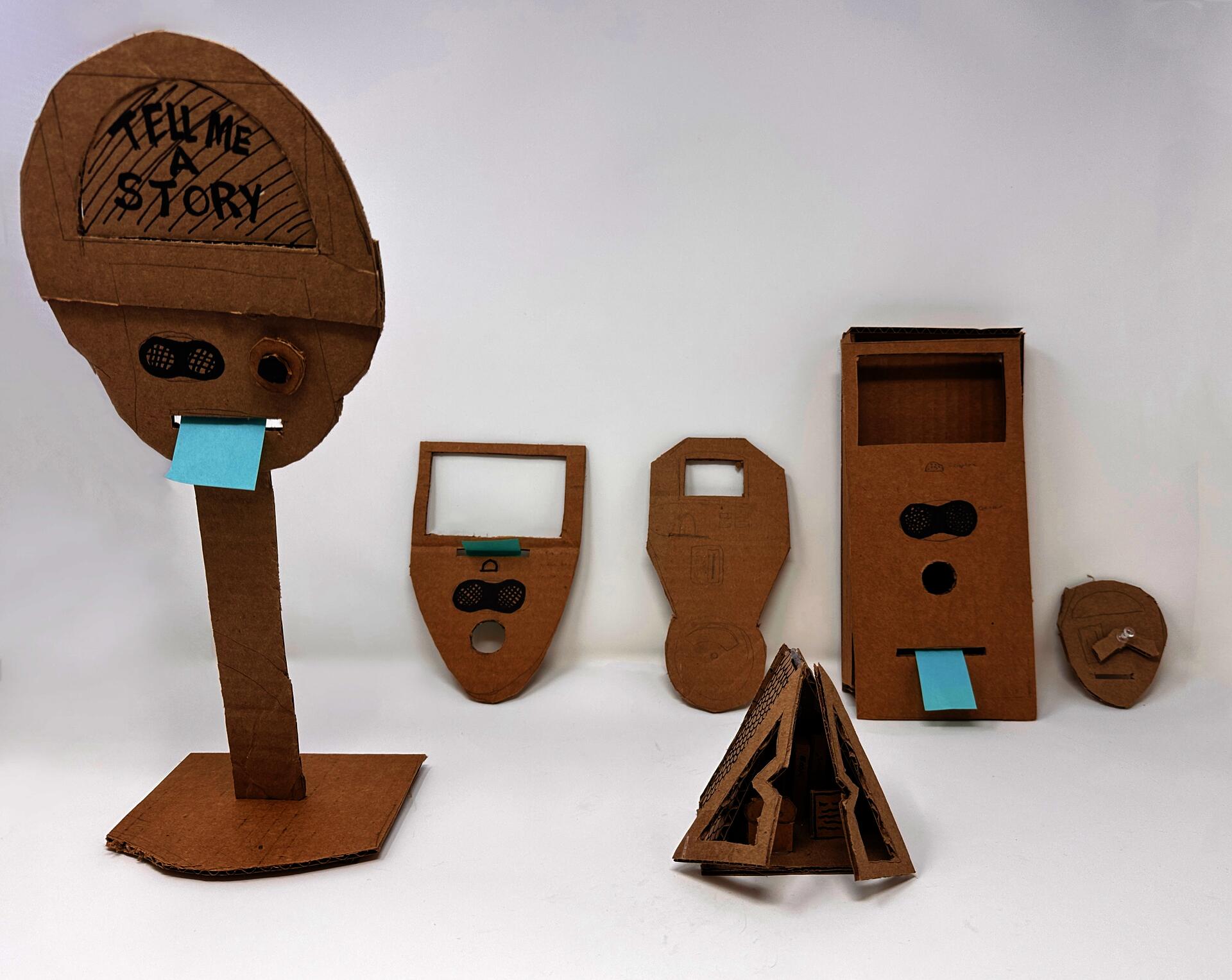 Six handmade cardboard prototypes of interactive objects, each with speaker grilles, a button hole, and a blue sticky-note "receipt" slot. The tallest reads "Tell Me A Story."