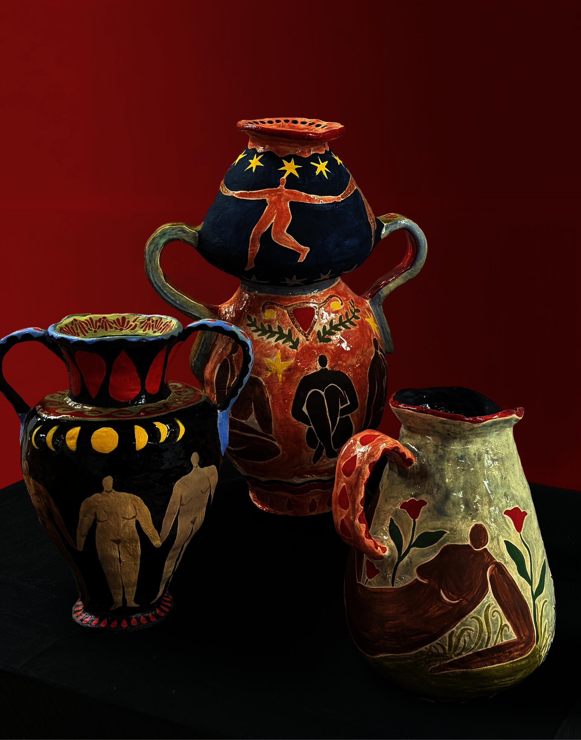 Three hand-painted ceramic vessels featuring figures, moons, stars, flowers, and menstrual imagery displayed on a black pedestal against a deep red background.