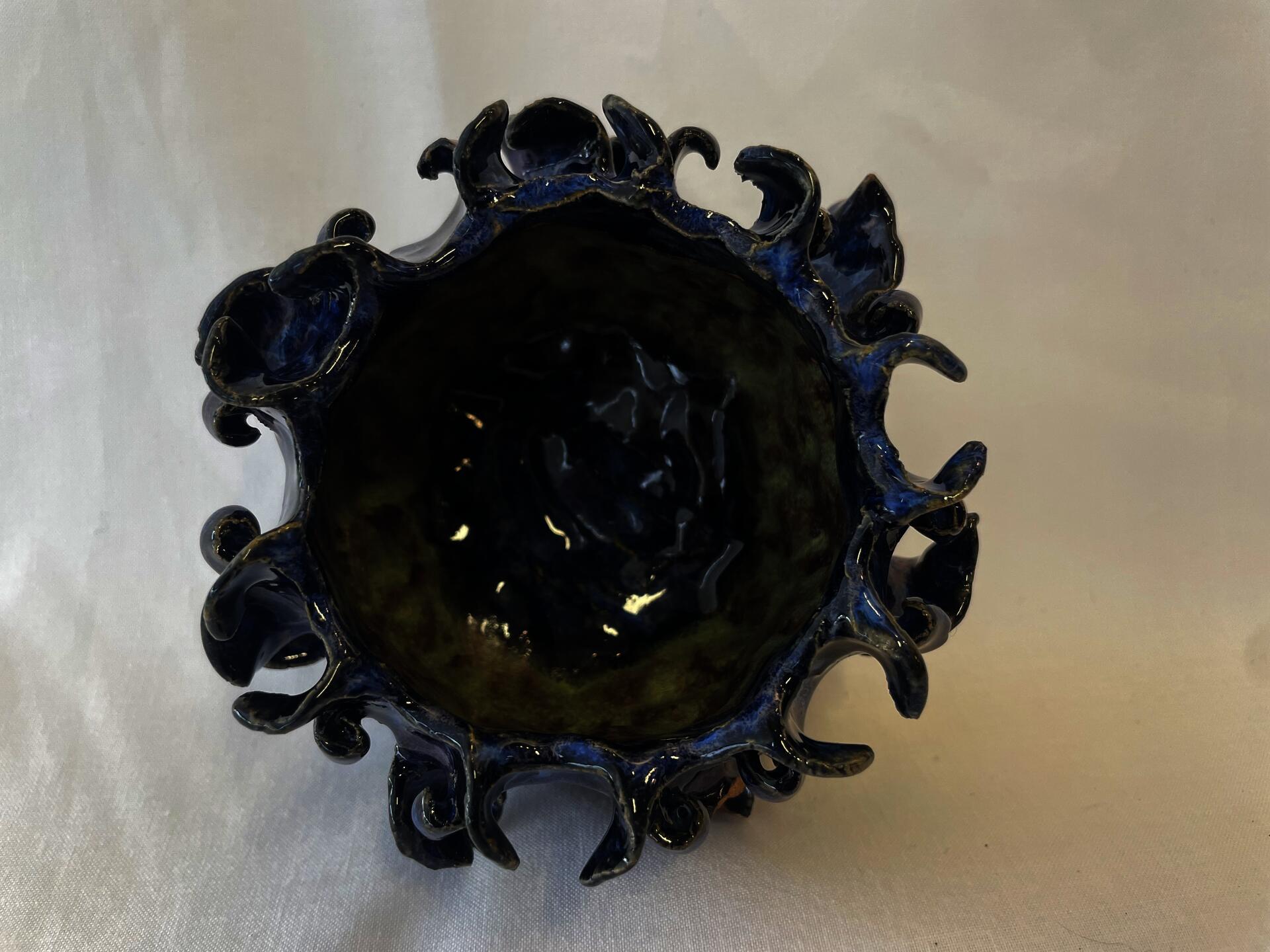 Ceramic bowl with leaf-like shapes surrounding the outside going in different pattern directions. Glaze colors are a  dark navy blue primarily with a green stripe on the inside.