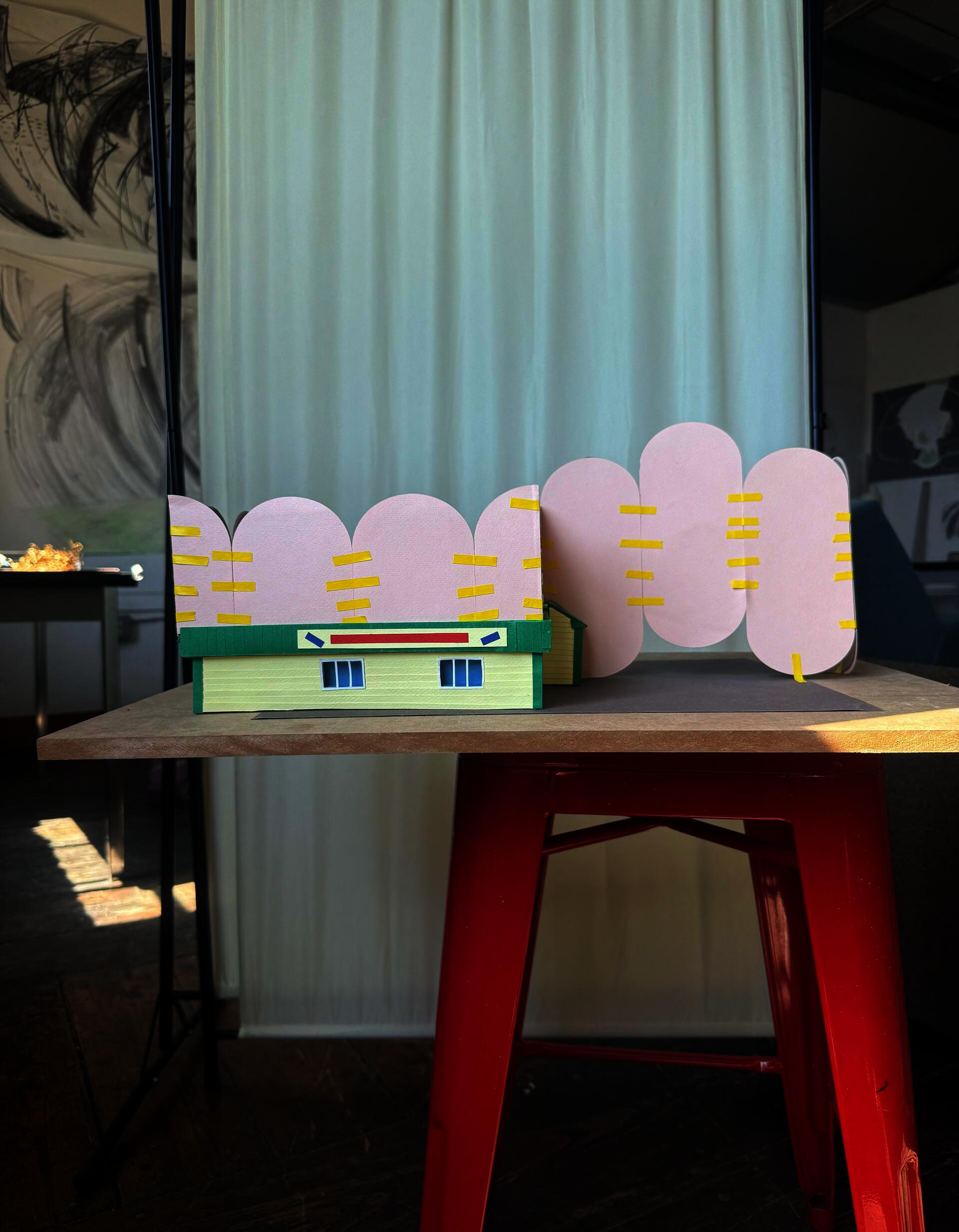 a paper model of a yellow laundromat with a green roof sits on a red stool in front of a cream curtain. Tall pink scalloped forms lay on top and adjacent to the building.