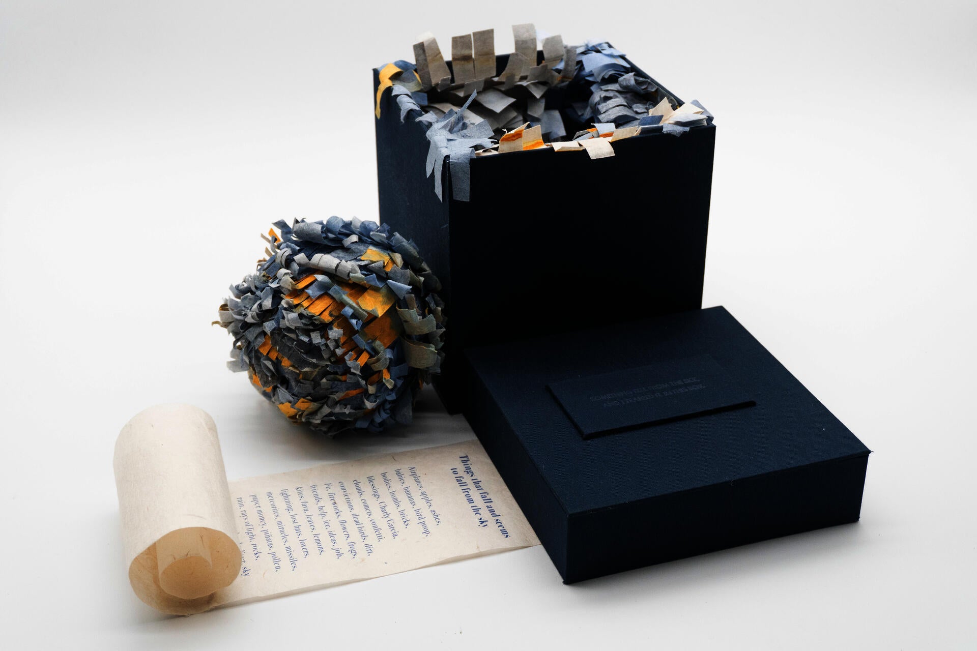 A small installation shaped like a book consists of a dark blue square box, a rolled handmade paper scroll, and a spherical handmade piñata covered in layered blue, gray, white, and yellow paper fragments.