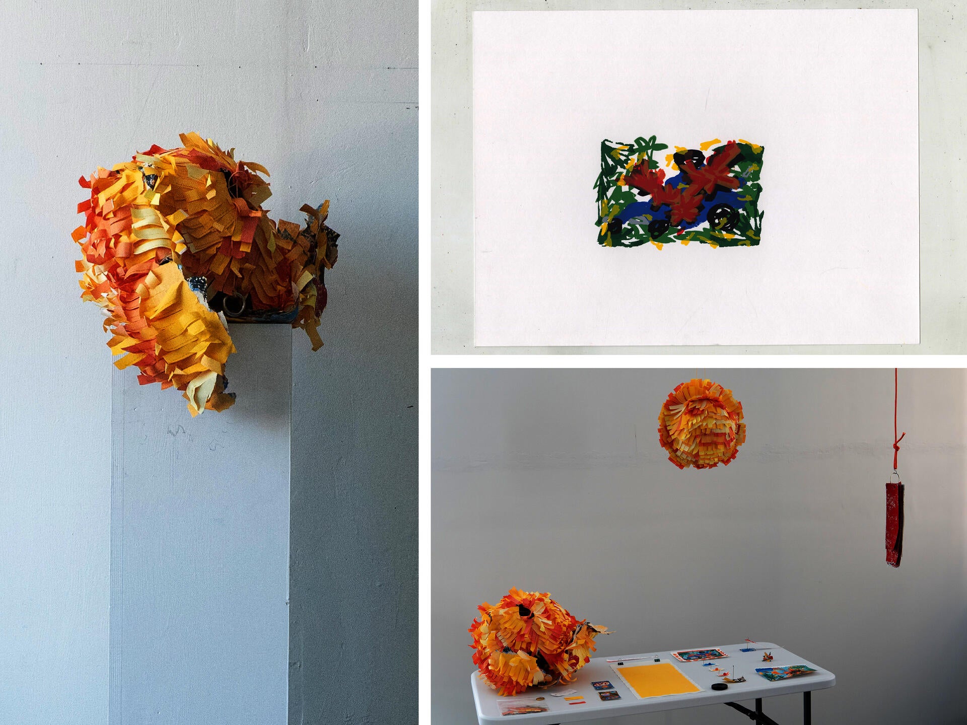 An installation features large piñatas made from layered strips of yellow, orange, and pink paper. One sculpture sits on a white pedestal, while another hangs from the ceiling above a white table displaying small printed images