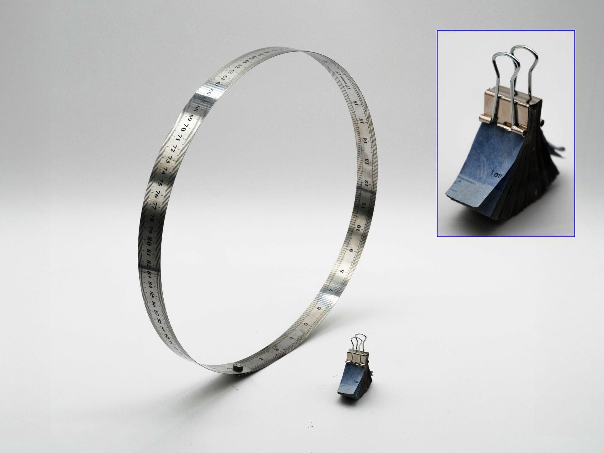A metallic ruler forms a large standing loop against a white background, bending away from its flat shape and held together with a small screw. Beside it sits a small stack of blue intaglio-printed paper pieces