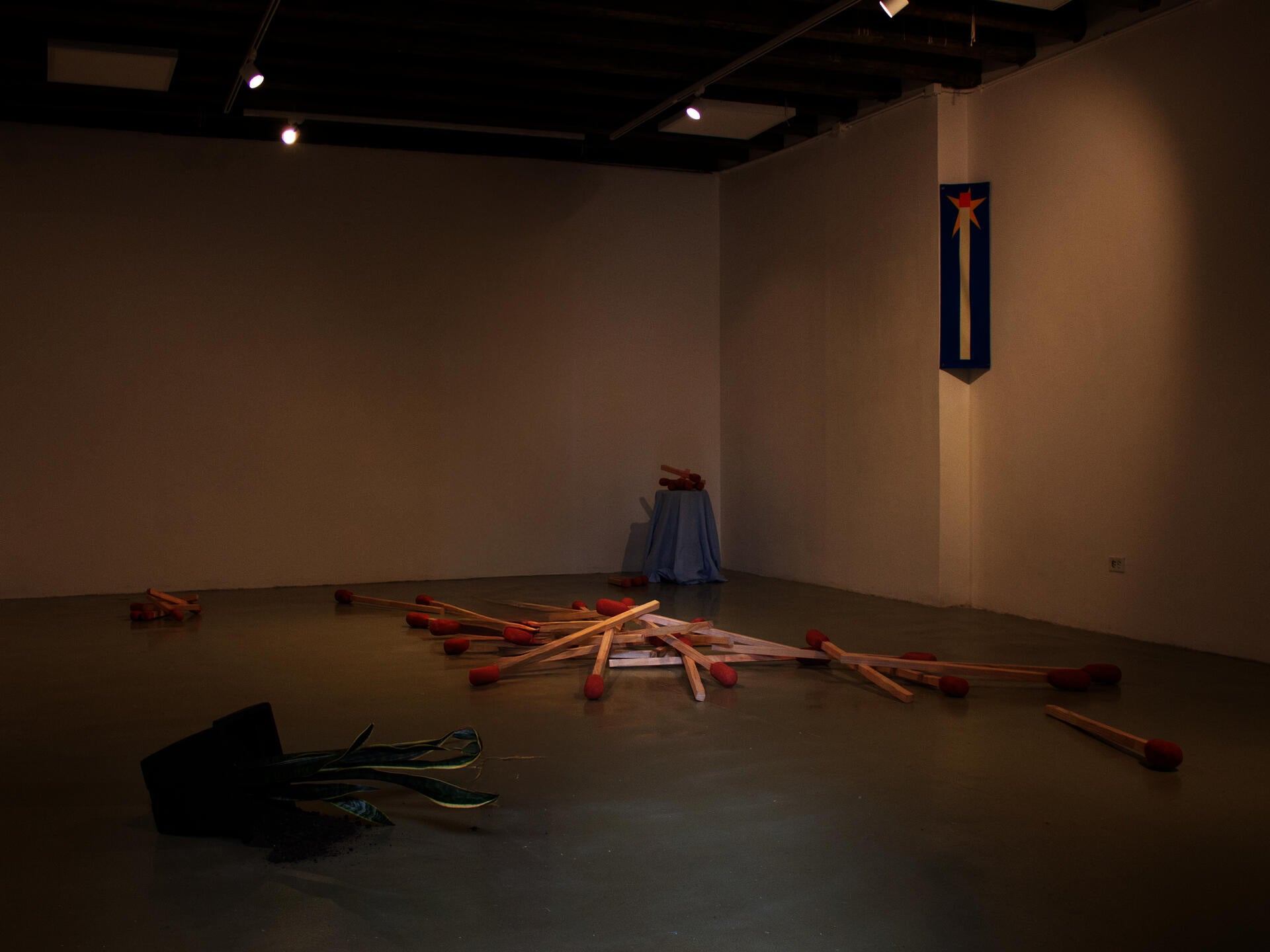 An installation fills a gallery floor with oversized wooden matches scattered in different directions. Each match has a large red tip and pale wooden shaft. In the foreground, a black flower pot lies tipped over with green paper leaves spilling out.