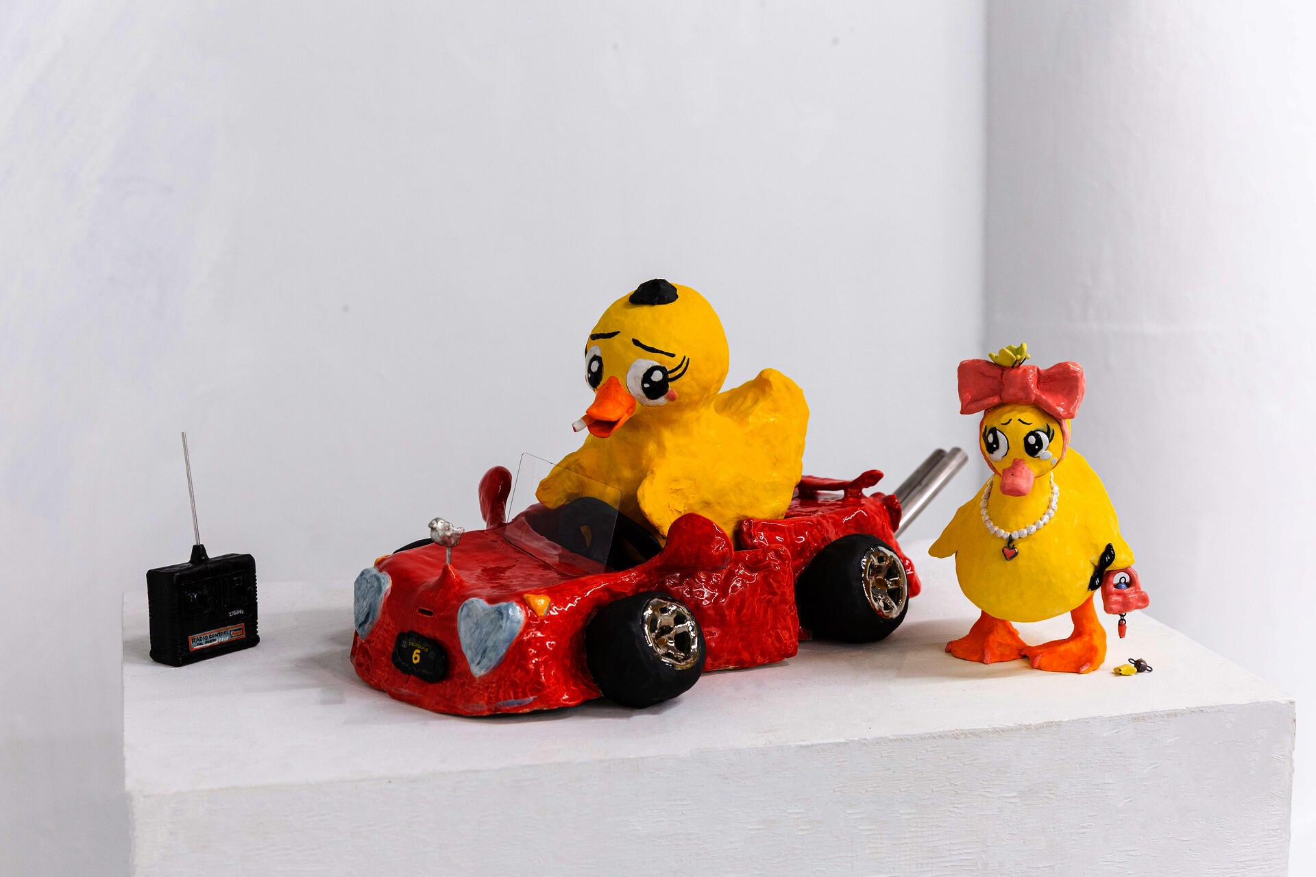 Ceramic sculpture of two ducks with human-like expressions in a red car. Glossy, playful yet uneasy, mixing childhood memory with subtle discomfort.