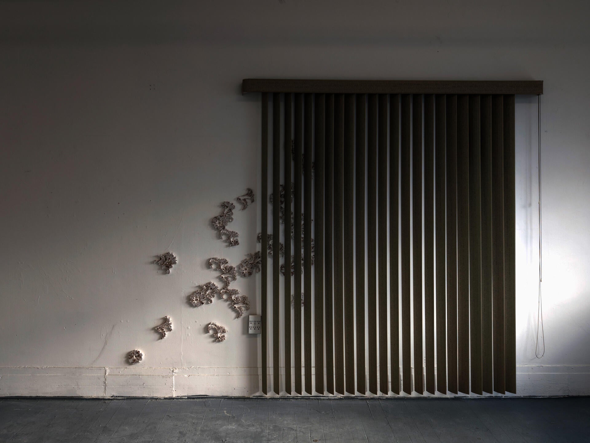 A set of vertical greenish blinds set on a white wall, a pattern of white flowers seep behind it.