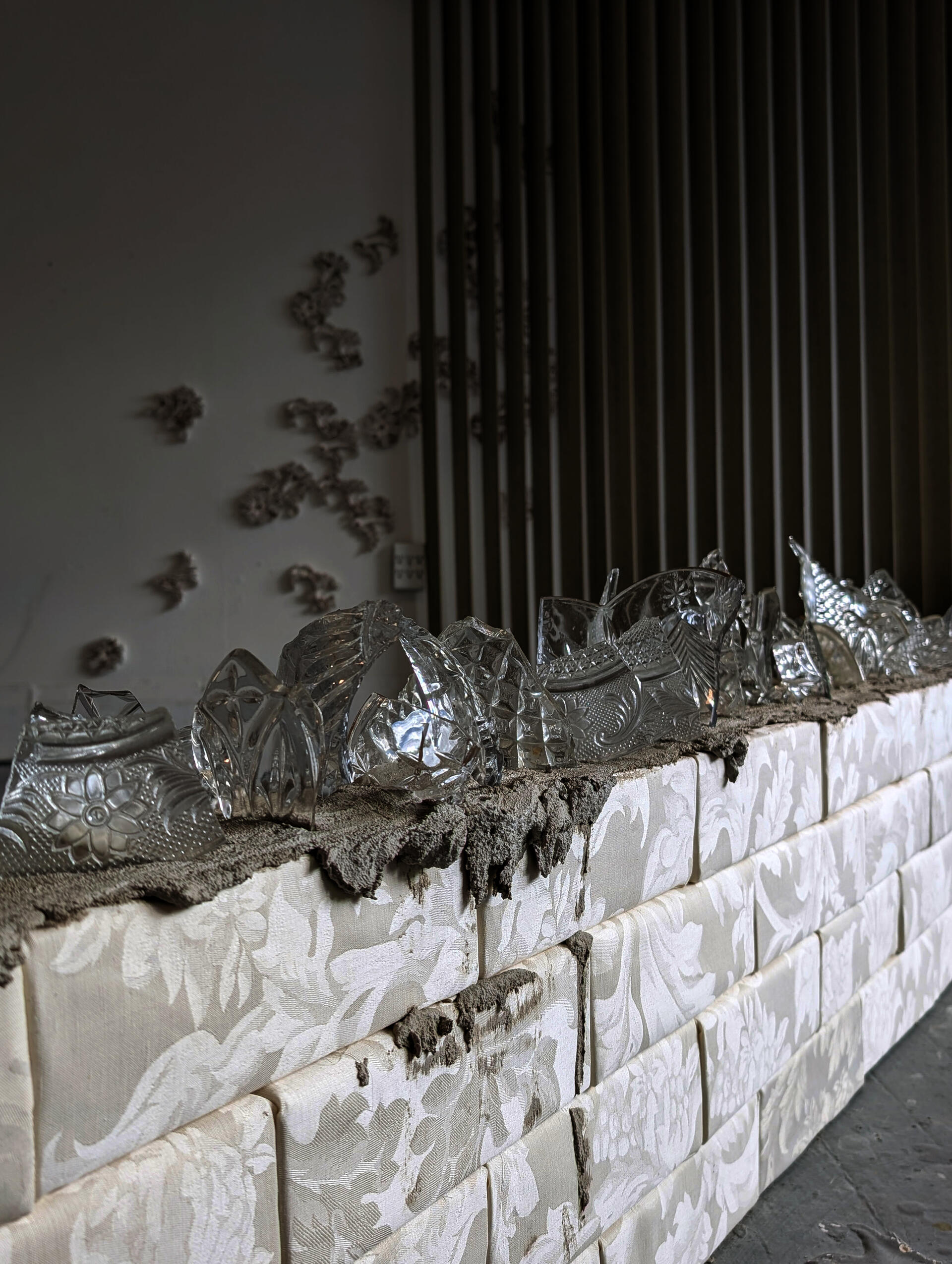 Broken antique glassware rests on a concrete bed atop a low wall built of upholstered, flower-patterned bricks.