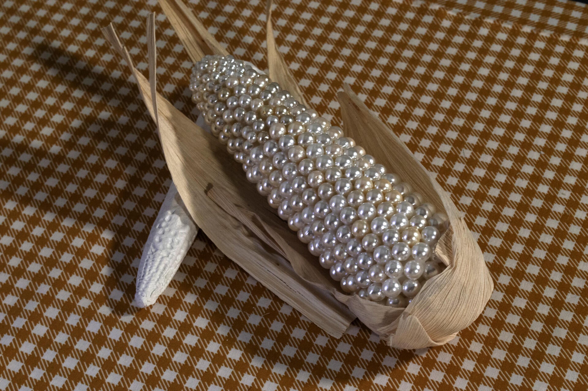 Fictional corn ear with faux pearls instead of kernels, rests on a gingham tablecloth. 