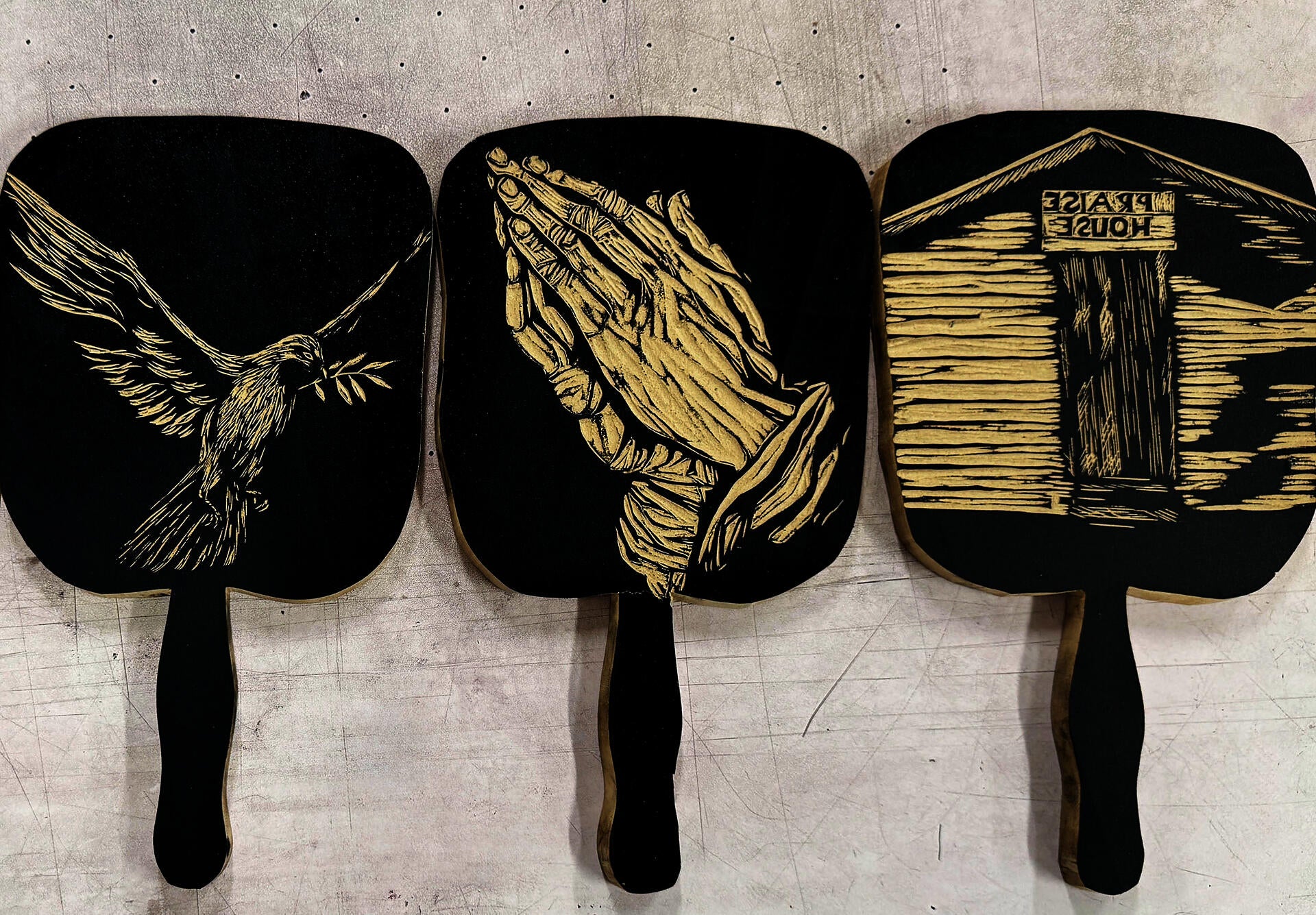 Three black and white wooden fans featuring Christian symbols: a dove with an olive branch, praying hands, and a miniature church that reads praise house. 
