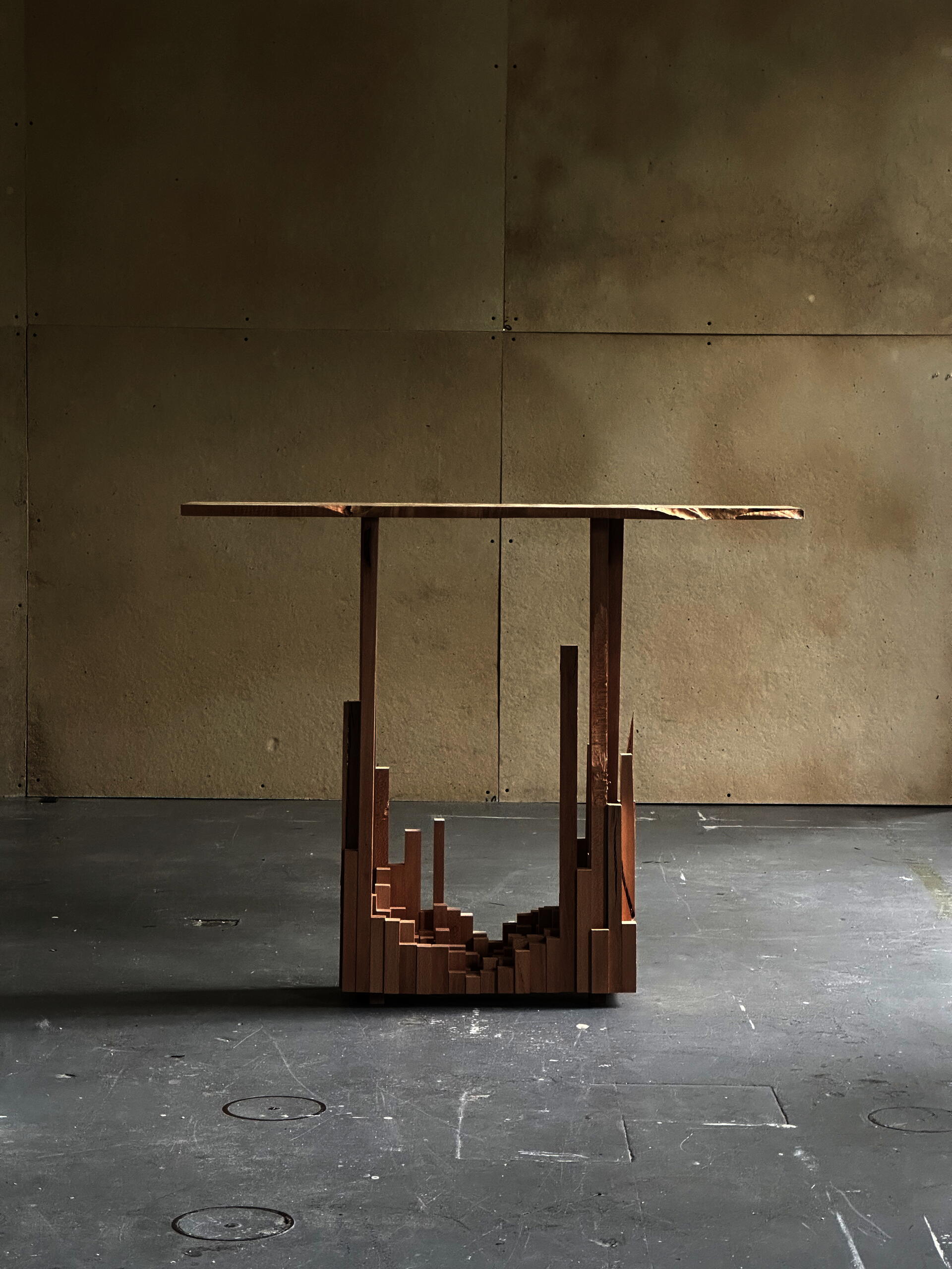 Minimalist wooden side table with a thin rectangular top supported by geometric vertical elements rising from clustered blocks. Warm wood tones create an architectural, skyline-like composition.