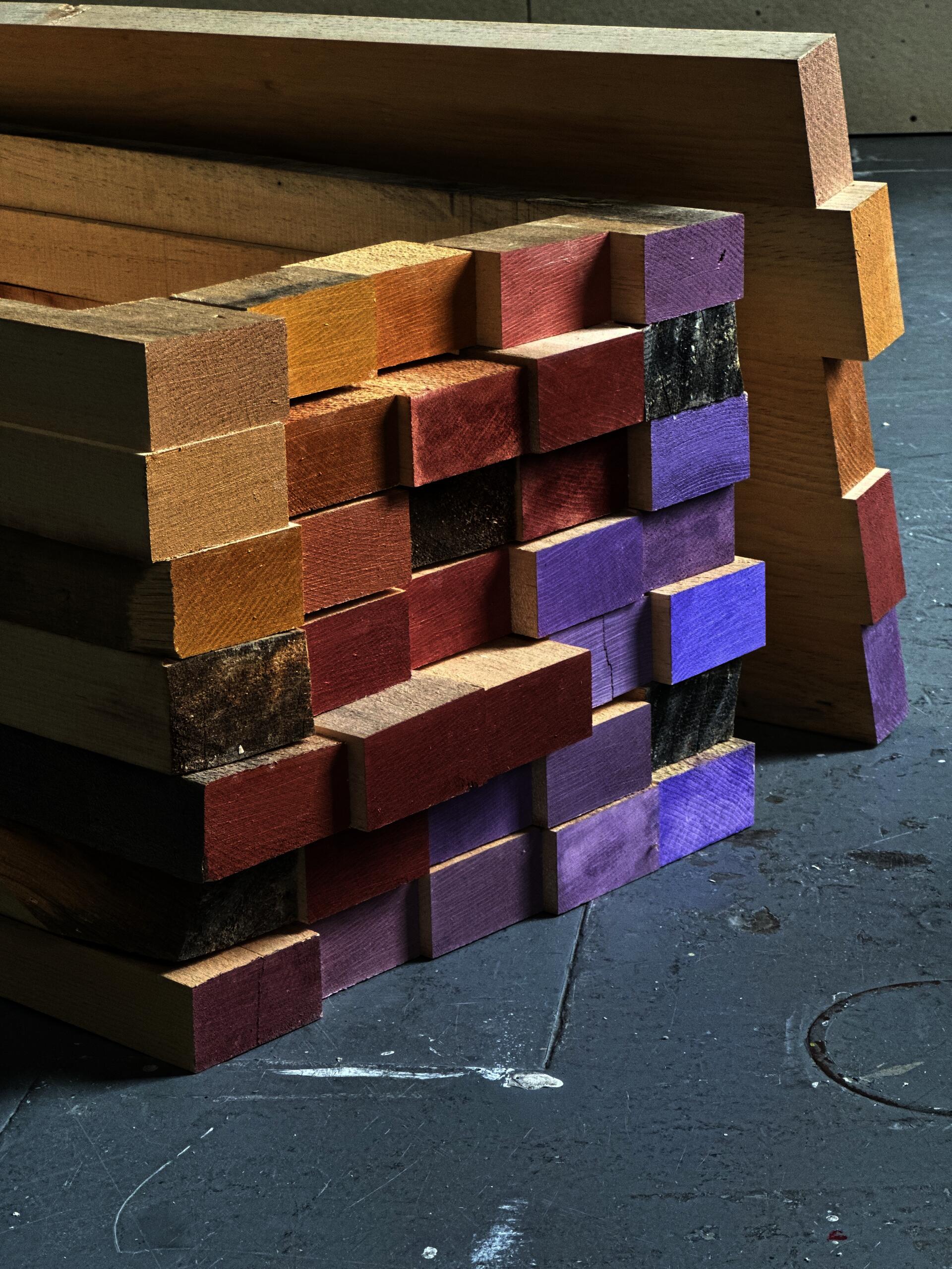 Stacked wooden blocks form a geometric table base with exposed end grain, varied wood tones, and painted surfaces. The layered construction emphasizes repetition, material contrast, improvisation, and the tension between rough construction and careful composition.