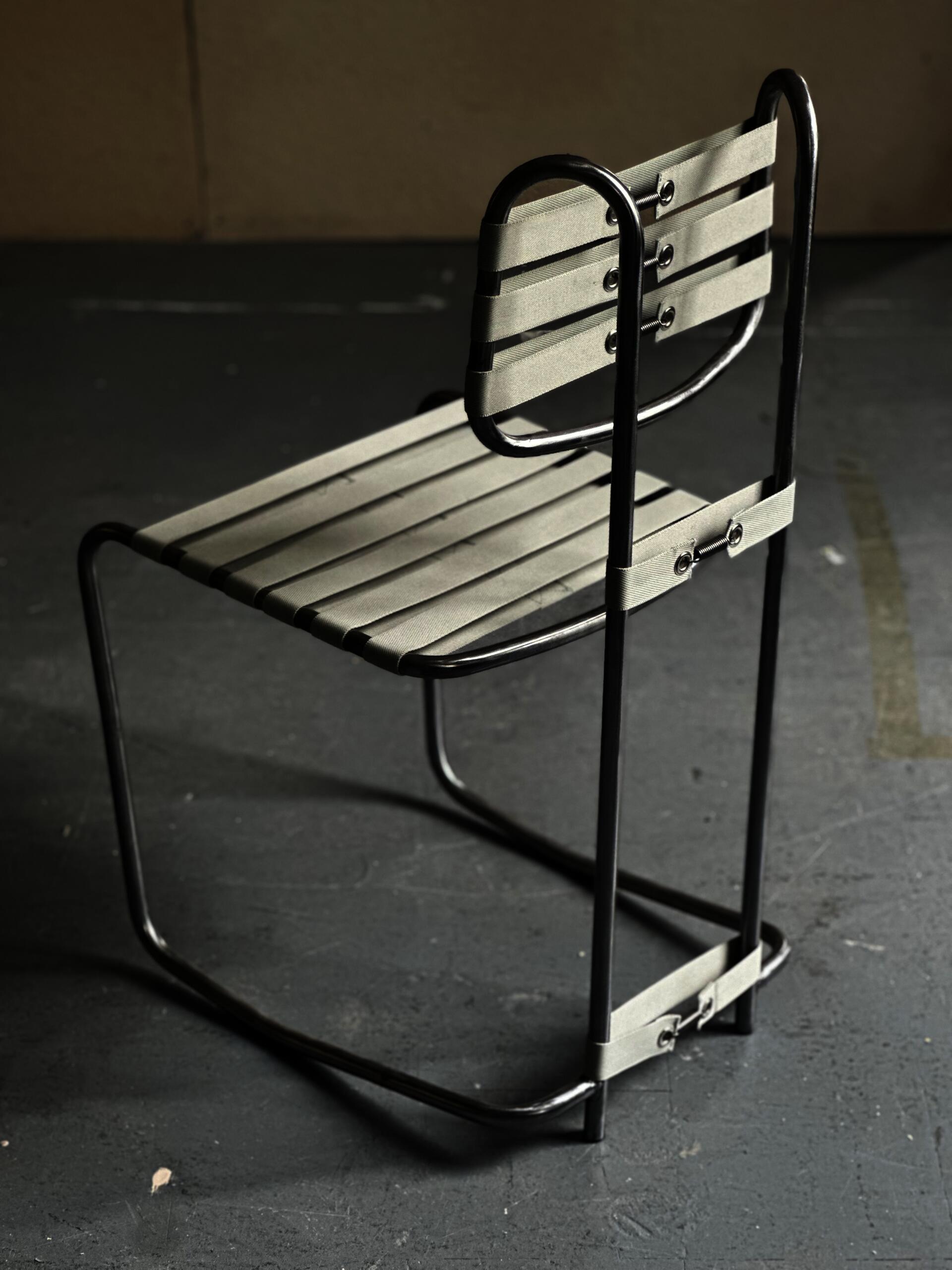 Cantilevered steel tube chair with tensioned nylon webbing seat and backrest. The continuous black frame contrasts with pale strapping, emphasizing flexibility, visible structure, active sitting, and the relationship between tension, support, and movement.