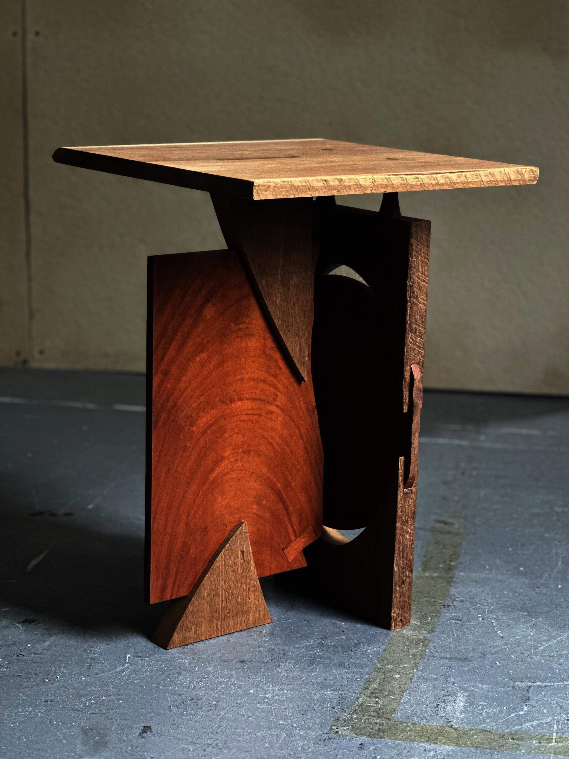 Small sculptural wooden table composed of interlocking geometric forms, contrasting wood tones, and exposed joinery.