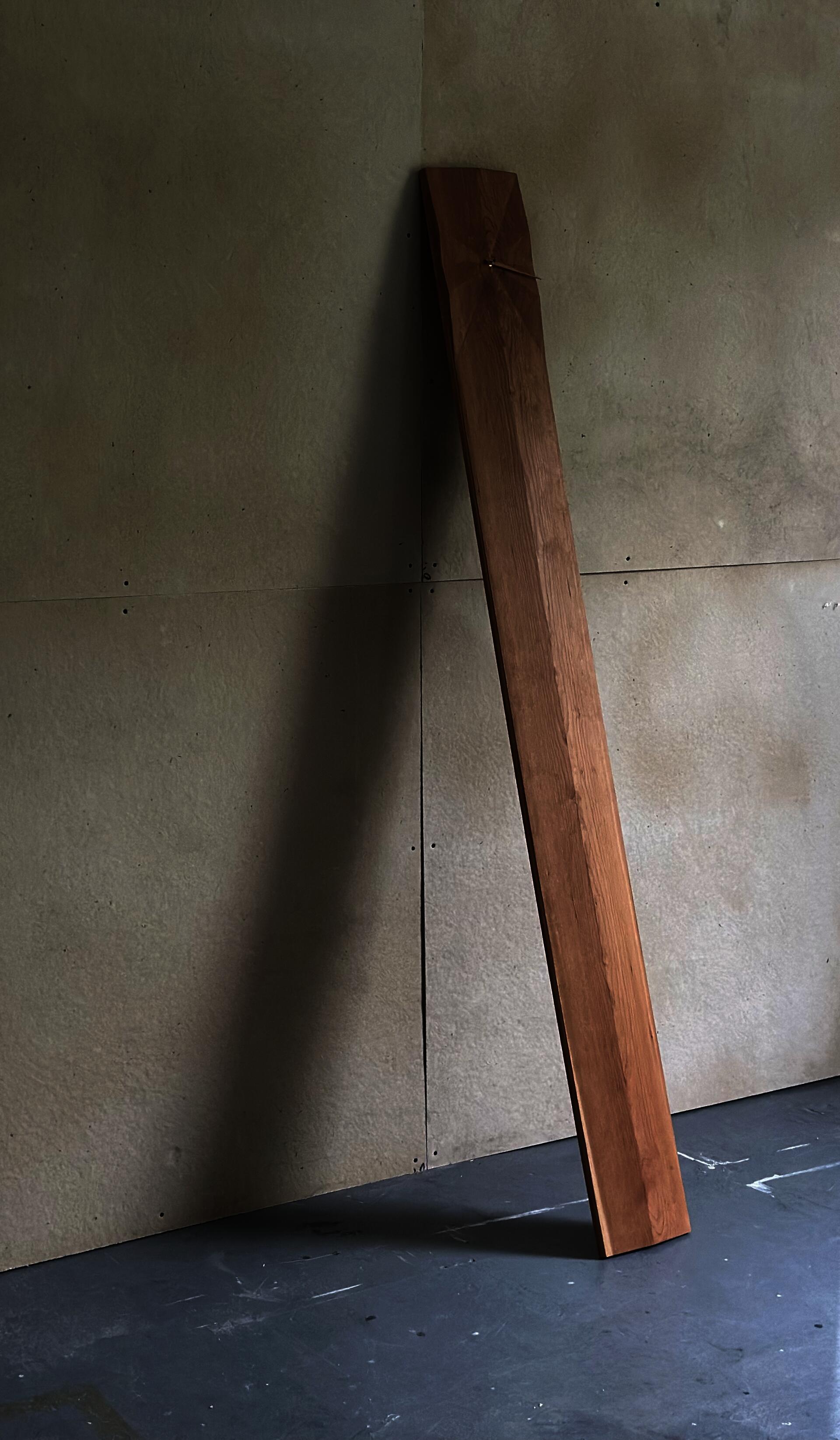 Minimal modernist cherry grandfather clock leaning casually against the wall, featuring a single hour hand and visible grain. The face is carved with a 12 radial facets.