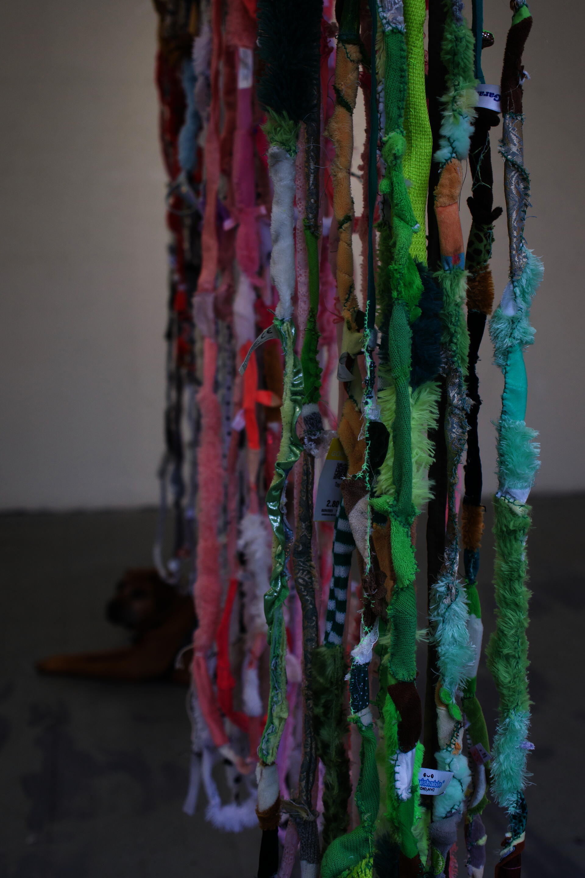 Close up profile of hanging stuffed animal rope. Green in focus with visible stitching, pink and red in the background, with loops as the bottom visible. Dog farther in the background.