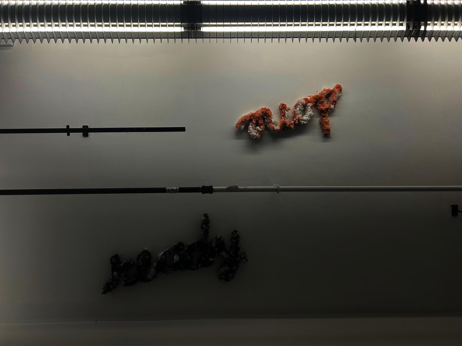 Ceiling shot, split by sprinkler piping and bordered by a florescent light. Cursive lettering-made from melted plastic shavings, stuffed animal fluff and pellets-is attached to the ceiling.