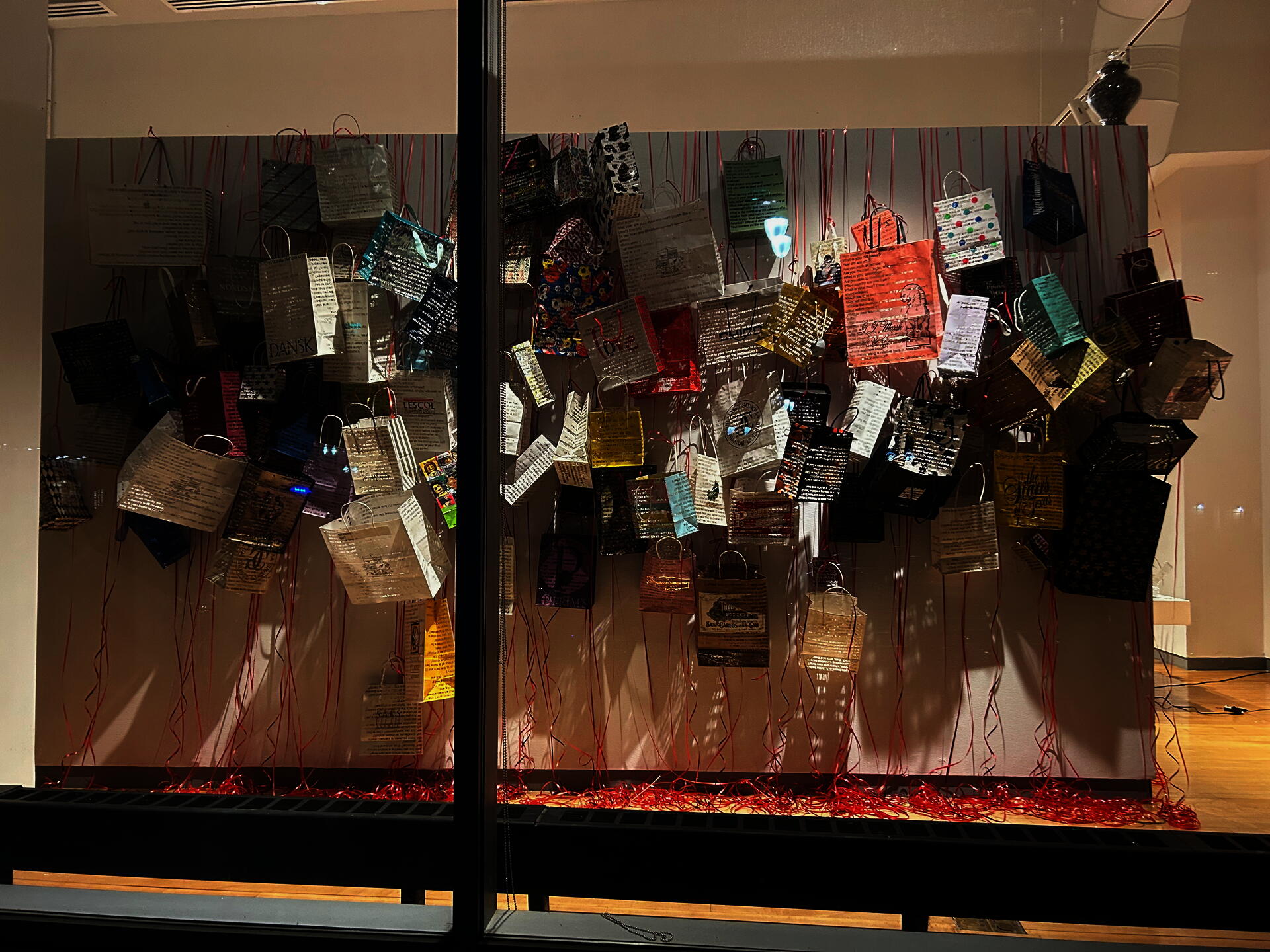 Window view of paper gift bags laser cut and hanging over a dividing wall. Variety of colors and sizes hanging from red party ribbon which spools and the floor beneath. Light casts shadows that show the cutouts.