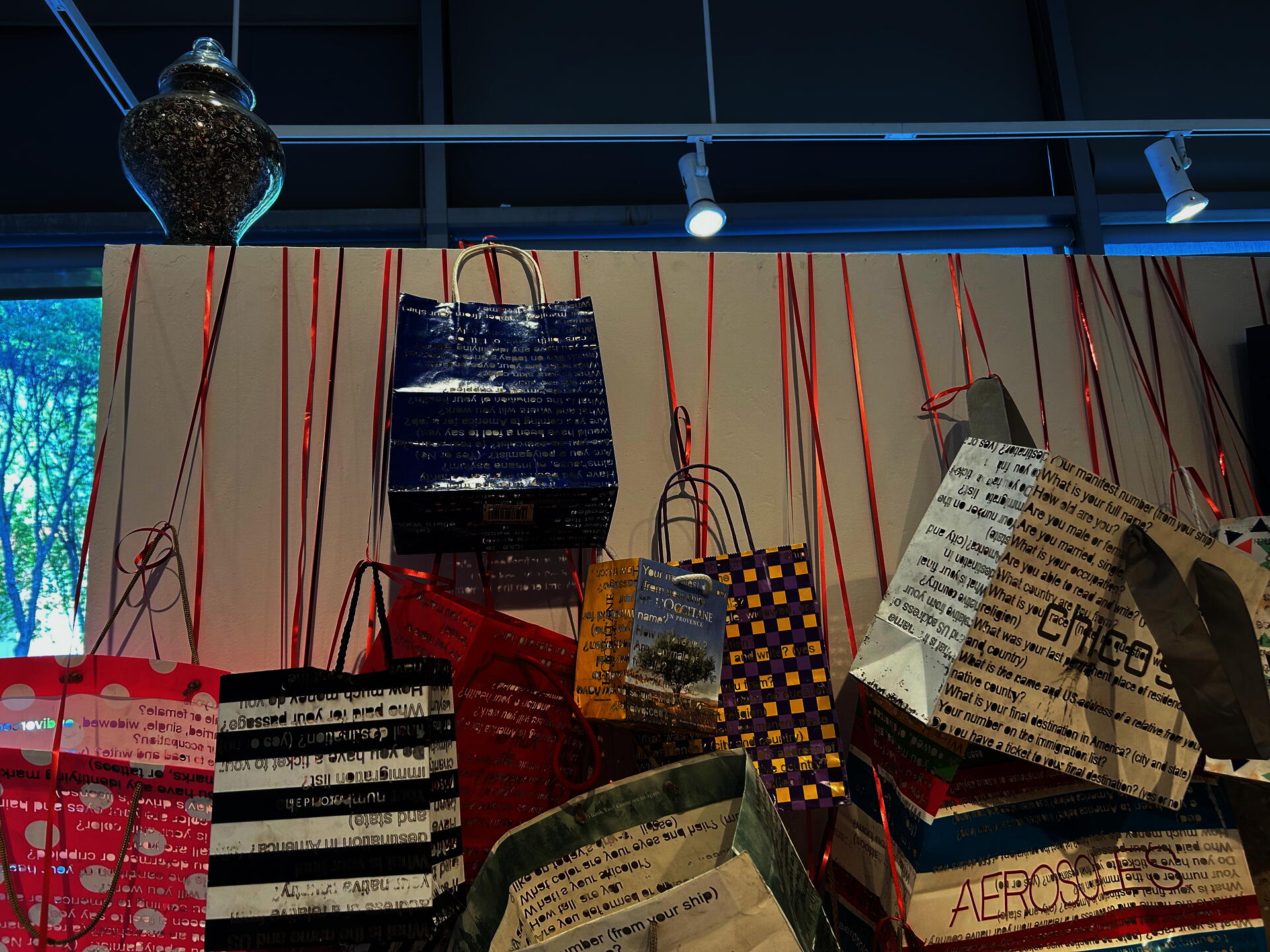 Close up of a few bags towards the top of the wall. Lettering is readable as the series of questions asked at Ellis Island. Glimpse of how the light casts through the cutouts. Letters in the