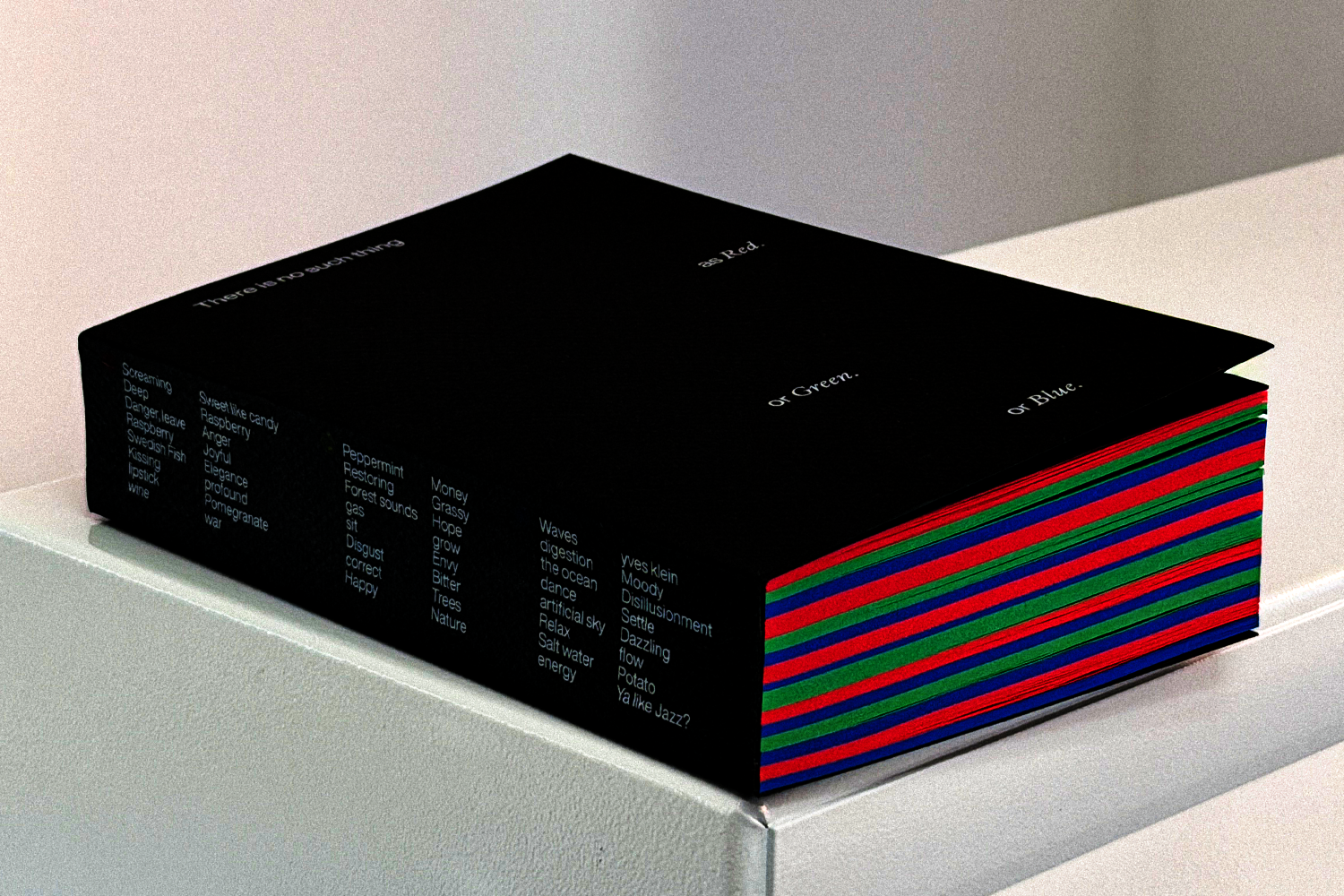 A 600-page book titled "There is no such thing as Red. or Green. or Blue." rests on a white pedestal. The spine lists participant survey responses. Page edges show alternating red, green, and blue stripes.