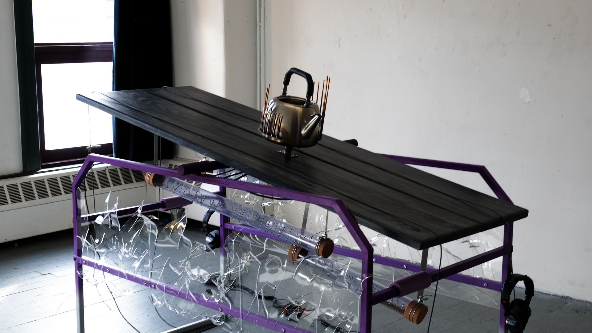 Purple steel table with kettle on wooden platform, ropes at both ends, clear acrylic sides showing rain sticks beneath, headphones attached.