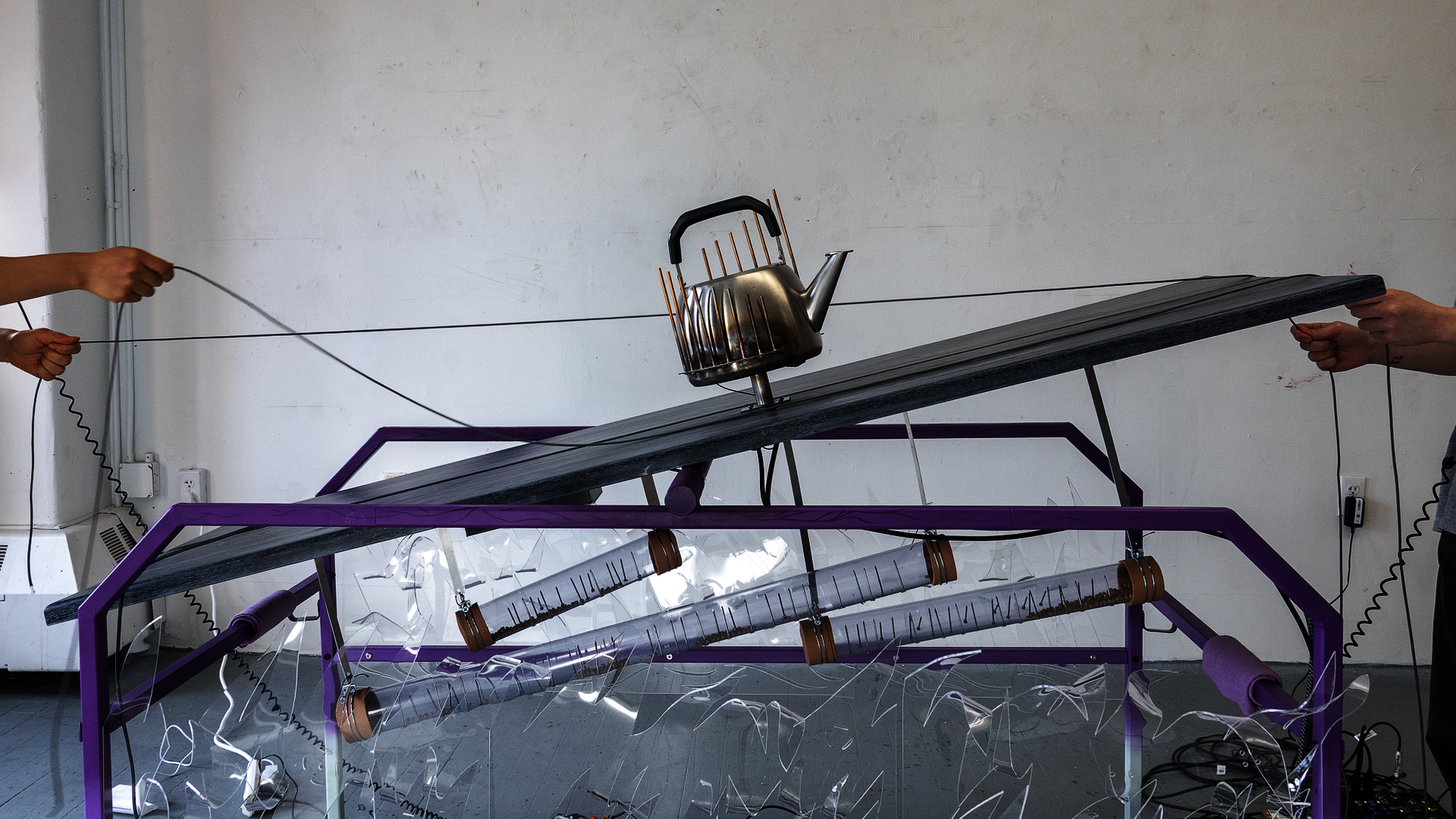 Two people pull ropes at opposite ends of a purple table, tilting a wooden platform with a waterphone-like kettle on top.