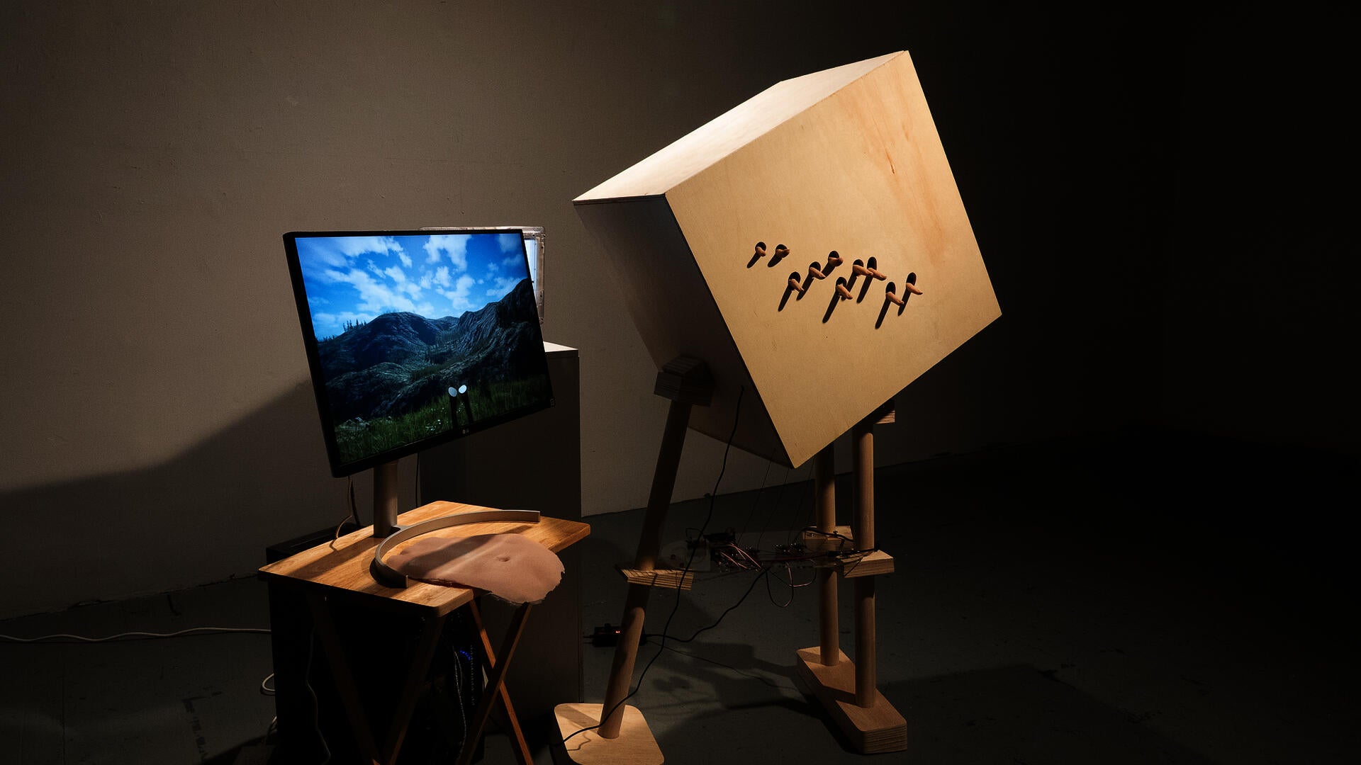 Installation with monitor showing digital landscape, silicone interface, and wooden structure with protruding fingers.